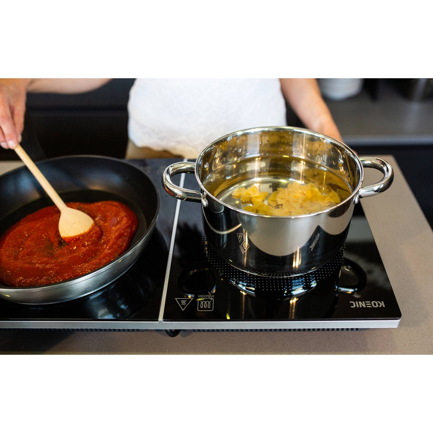 Una persona sta cucinando. Salsa di pomodoro in padella, pasta in una pentola con acqua. Entrambi sono su un piano cottura a induzione nero.