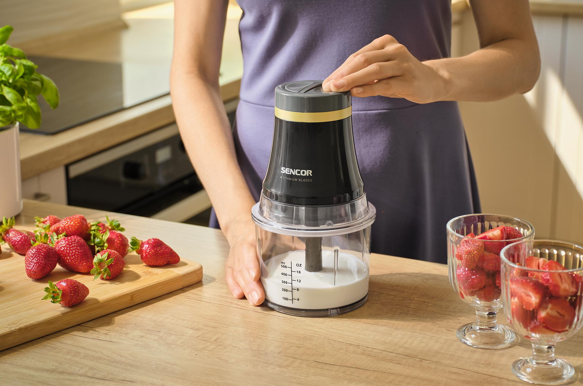 Egy nő turmixgépet használ smoothie készítéséhez. Eper és eperdarabokkal teli poharak vannak az asztalon.
