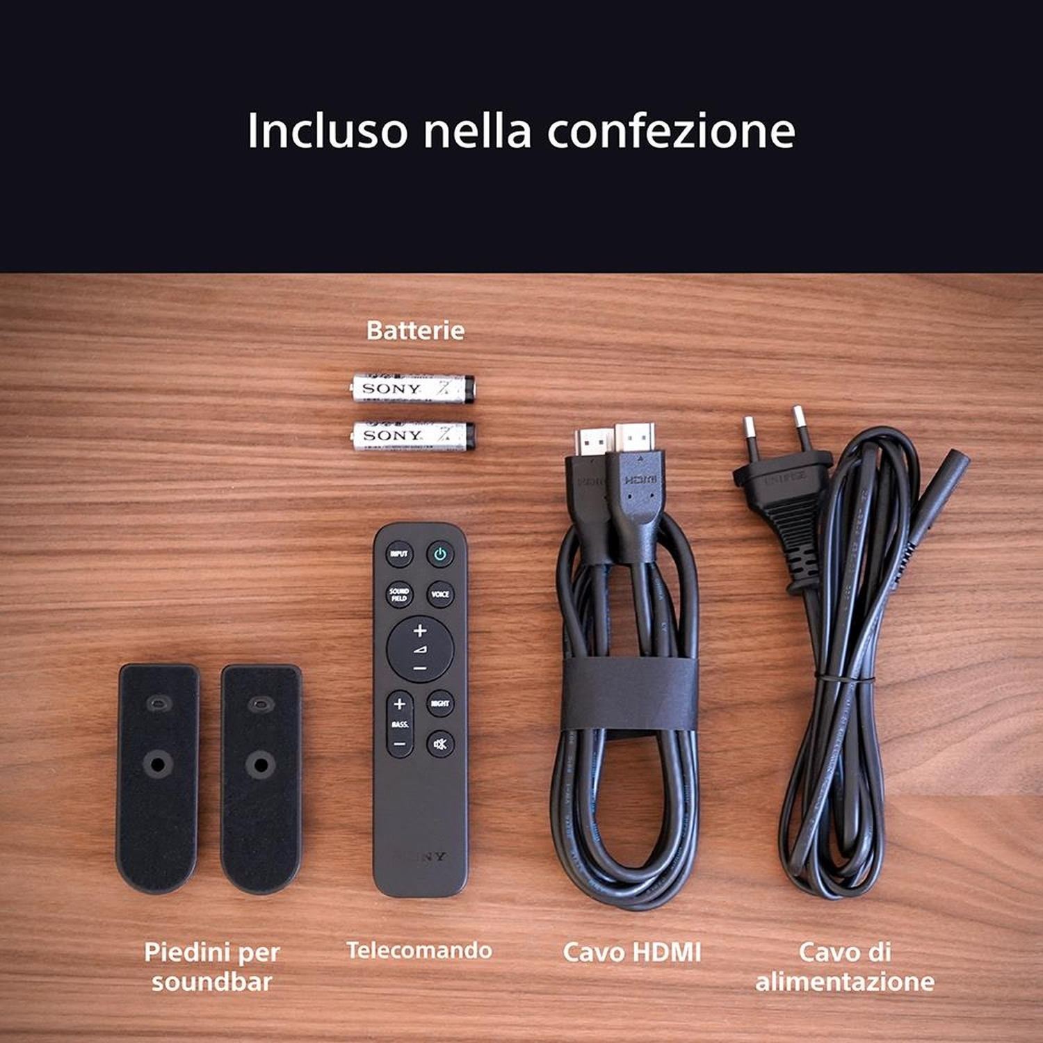 L'immagine mostra il contenuto di una scatola. Include batterie, telecomando, piedini soundbar, cavo HDMI e cavo di alimentazione, su superficie in legno.