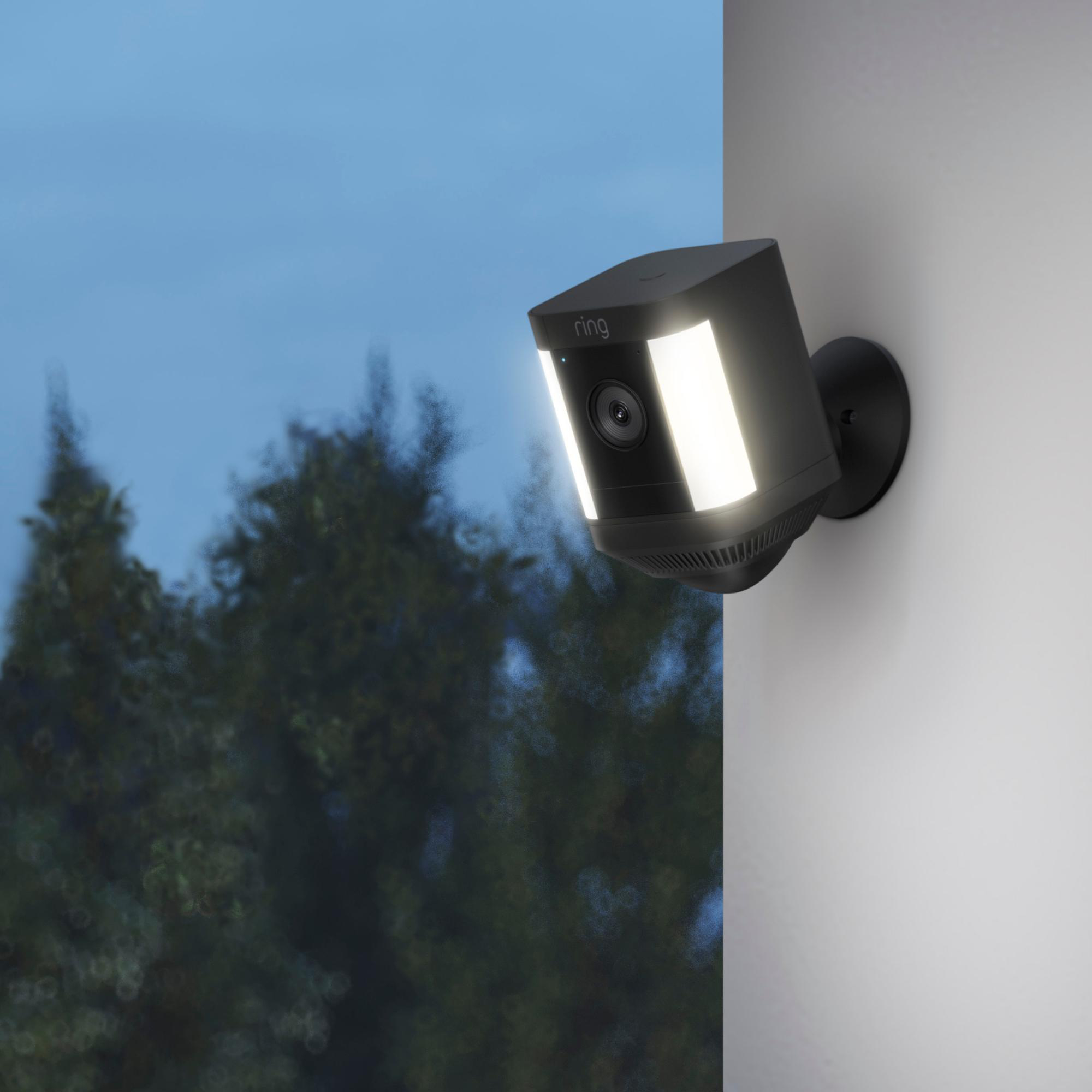 Eine schwarze Ring-Überwachungskamera mit Lichtern, die an einer weißen Wand montiert ist. Blauer Himmel und Bäume im Hintergrund.