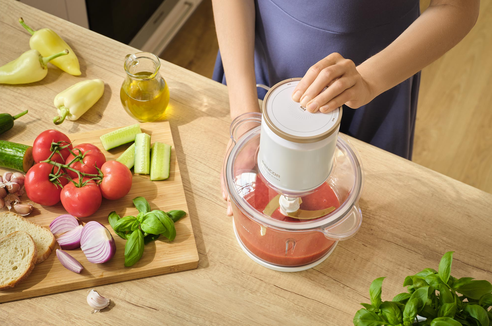 Egy személy egy konyhai robotgépet használ. Zöldségek, mint a paradicsom, uborka és paprika egy vágódeszkán.