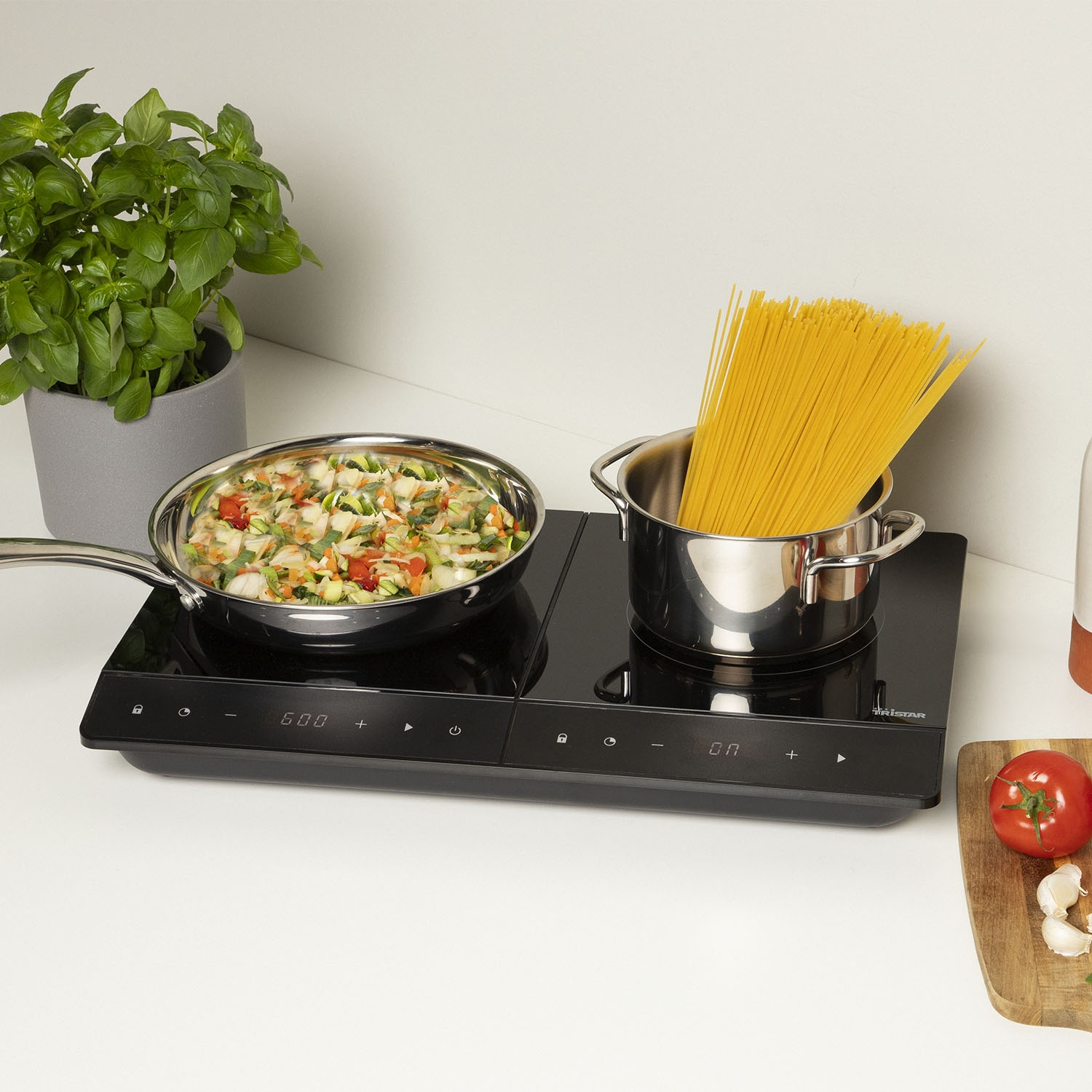 Un piano cottura con una padella di verdure e una pentola di spaghetti. Una pianta in vaso è accanto, e un pomodoro con l'aglio.