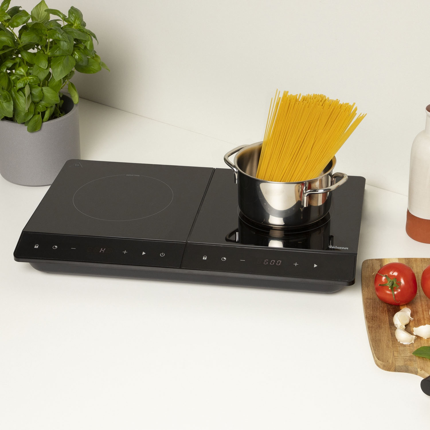 Un piano cottura a induzione nero con una pentola di spaghetti, accanto a pomodori, aglio e una pianta di basilico.