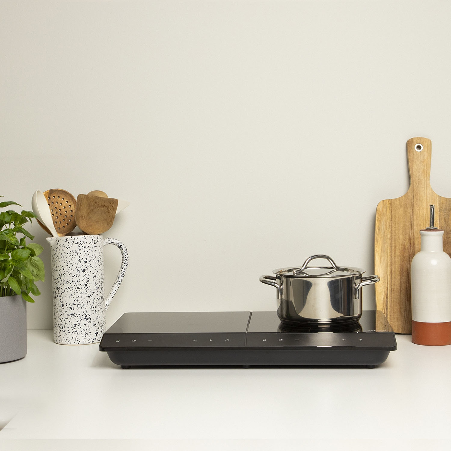 Una scena di cucina con piano cottura nero, pentola in acciaio inox, tagliere in legno e utensili in un vaso maculato.