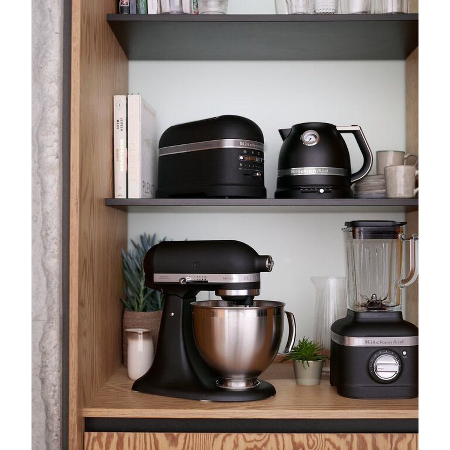 Un mobile da cucina con elettrodomestici neri: un tostapane, un bollitore, un mixer e un frullatore. Sono visibili anche libri e piccole piante.