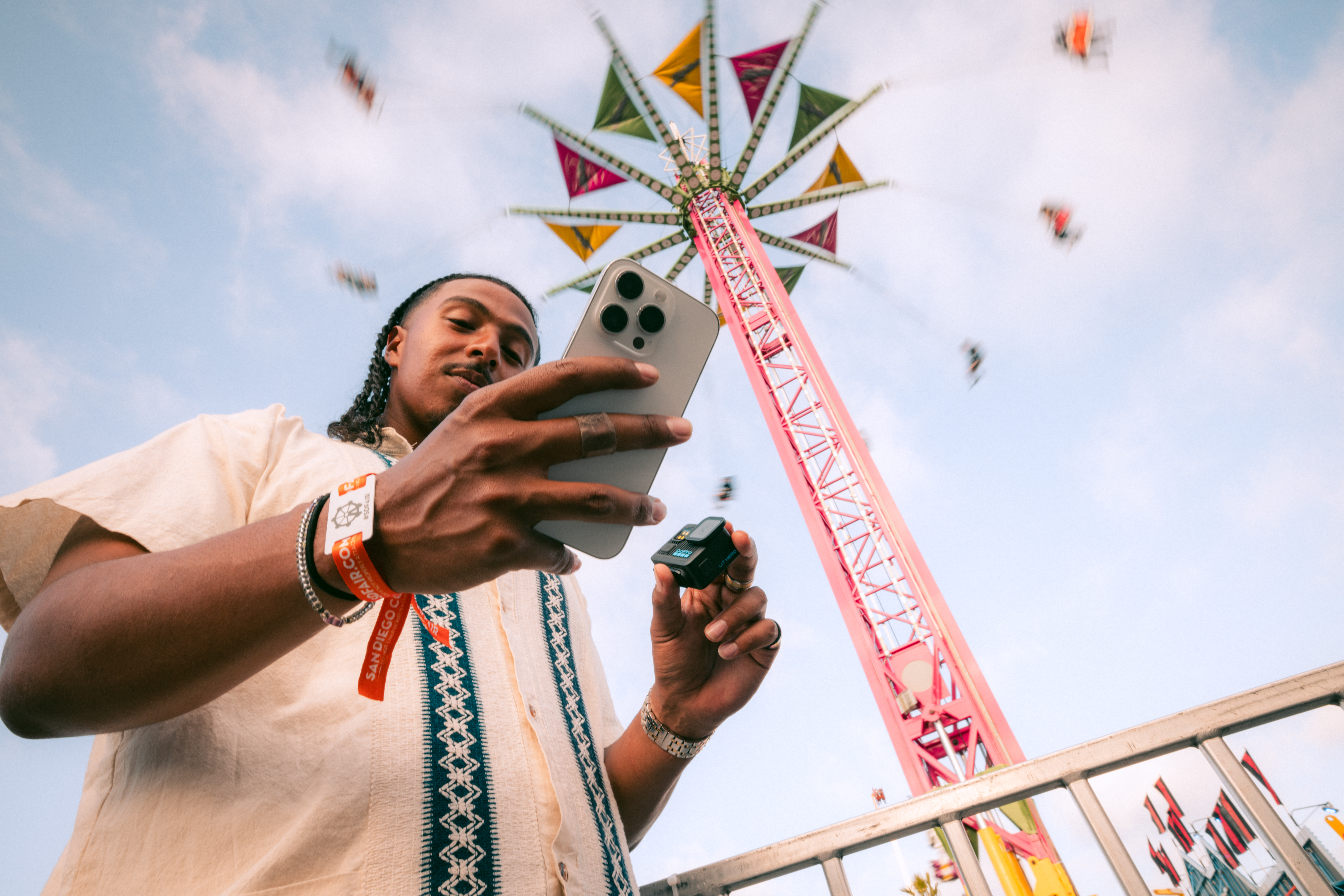 Mann hält Telefon und Kamera auf dem Jahrmarkt. Riesenrad im Hintergrund, blauer Himmel. Er trägt ein Hemd und Armbänder.