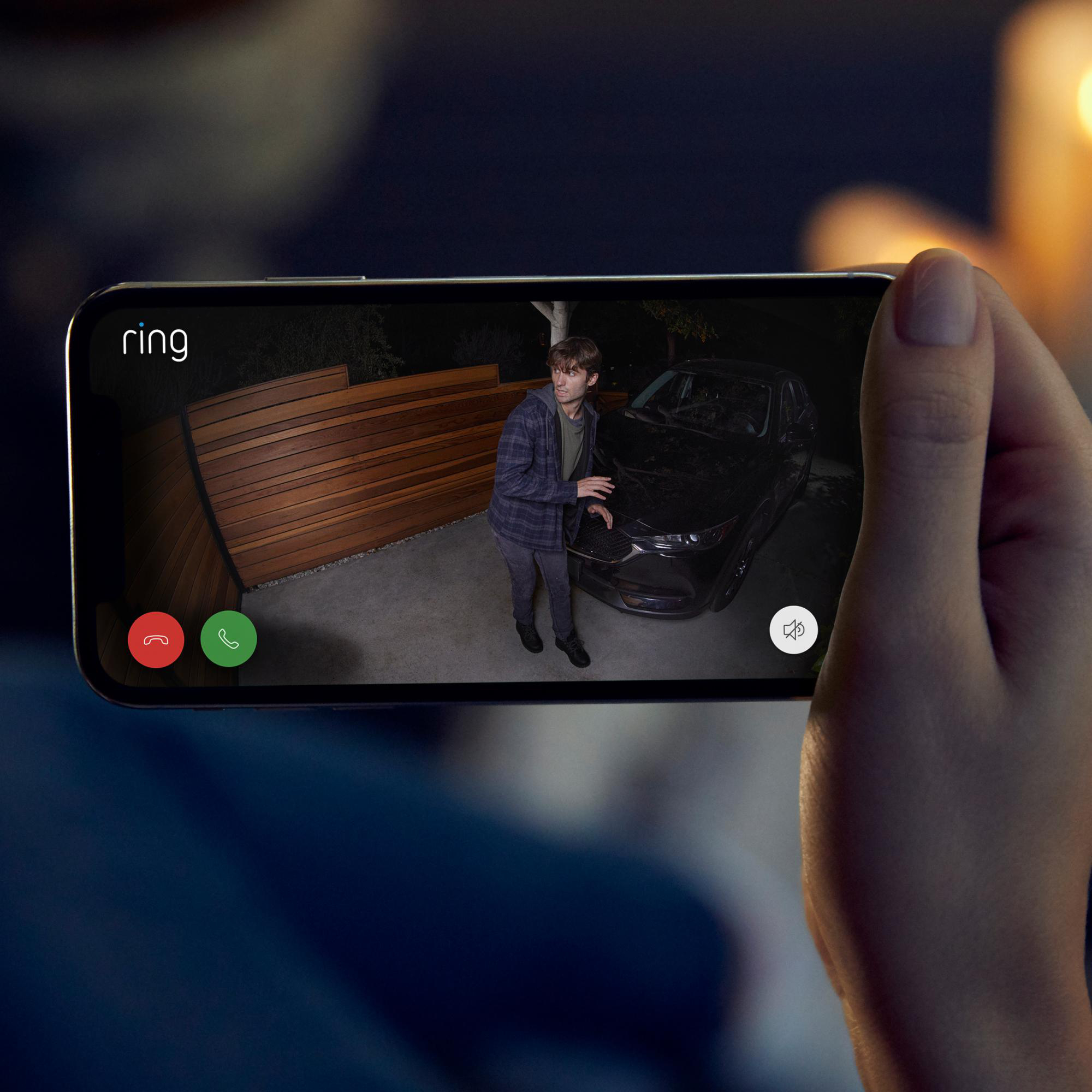 Eine Person hält ein Telefon, das eine Ring-Kameraansicht einer Person in der Nähe eines Autos bei Nacht zeigt.