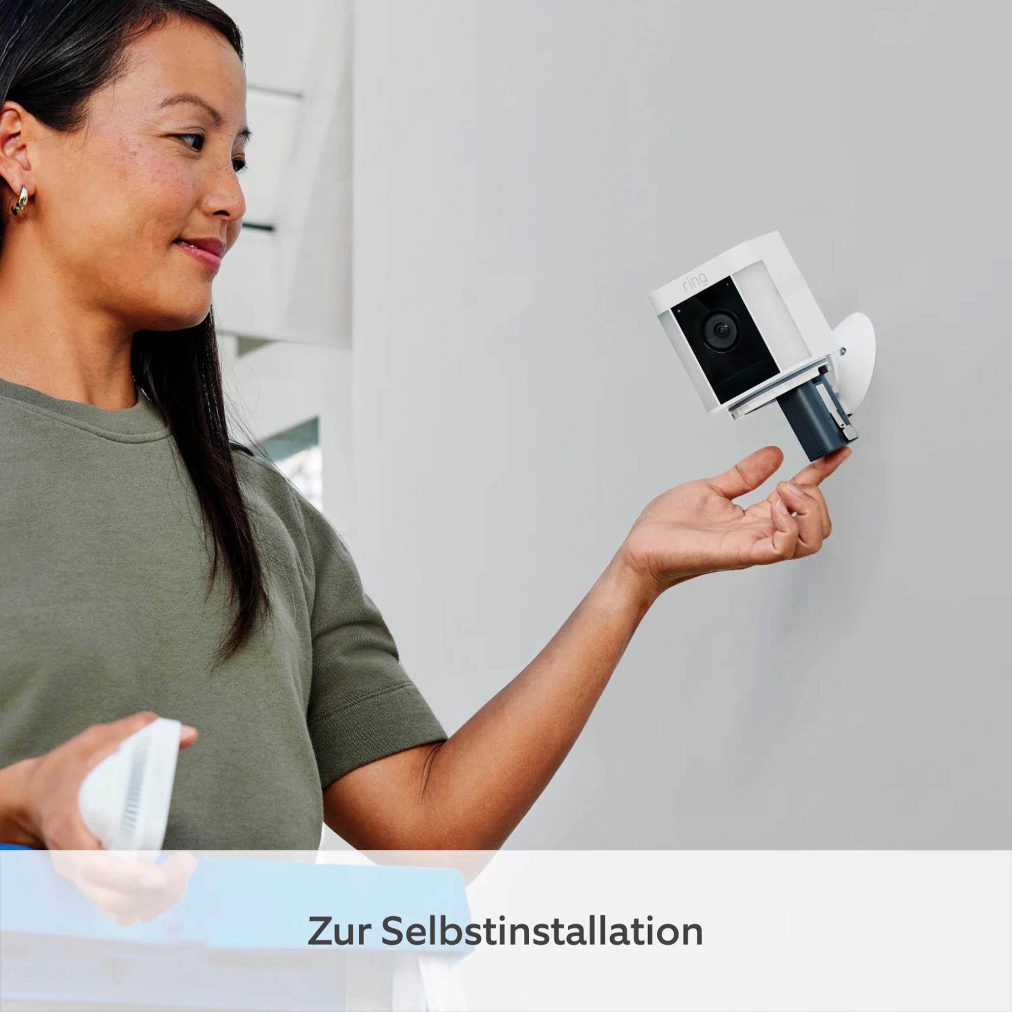 Frau hält eine Sicherheitskamera, lächelt. Kamera an der Wand montiert. Text unten: 'Zur Selbstinstallation'.