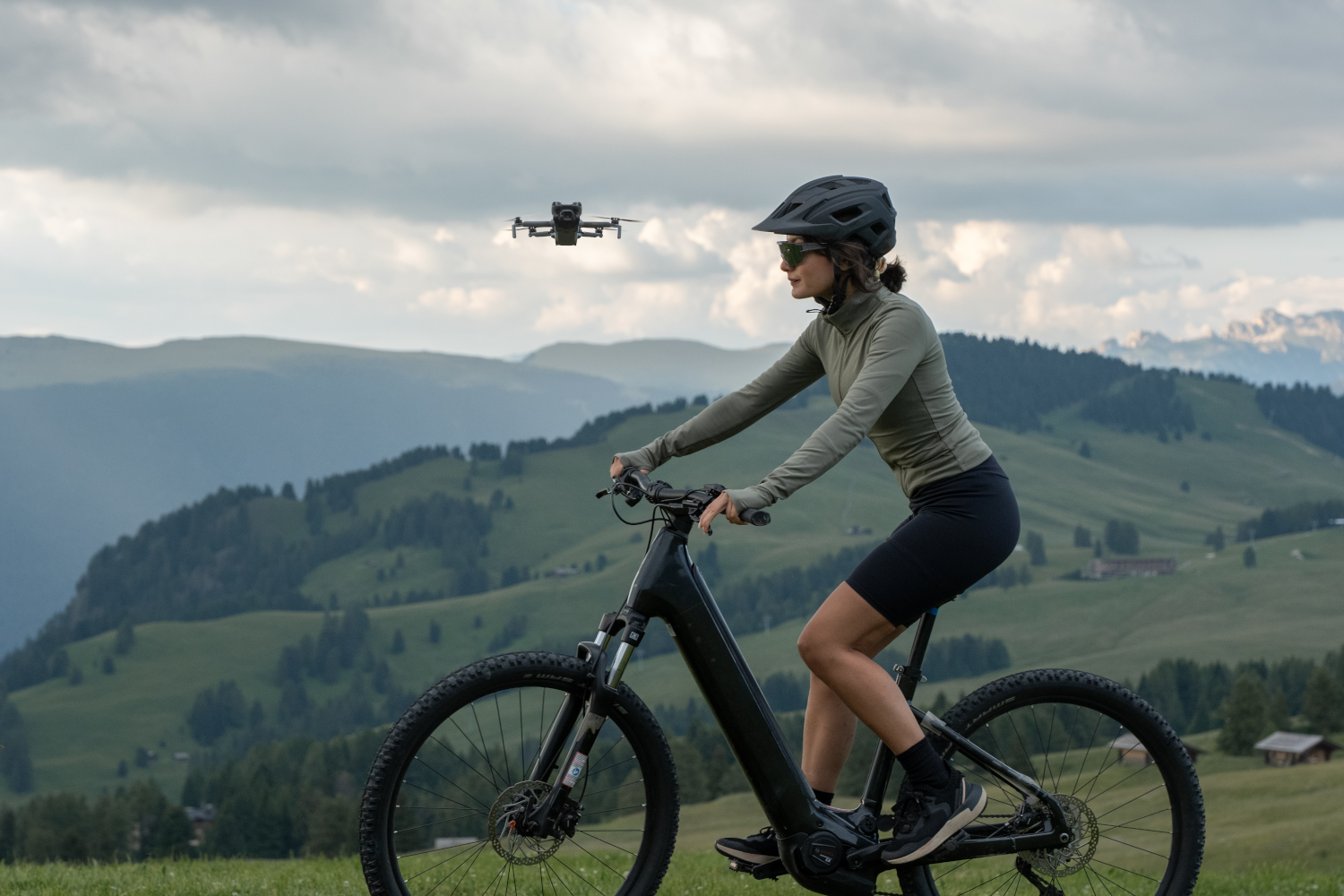 Una donna in mountain bike su una collina erbosa, con un drone che vola sopra. Montagne e cielo sullo sfondo.