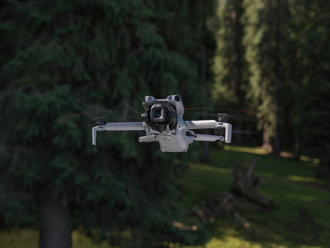Un drone grigio, DJI Mini 3 Pro, è in volo. Lo sfondo presenta alberi e un paesaggio verde sfocato.