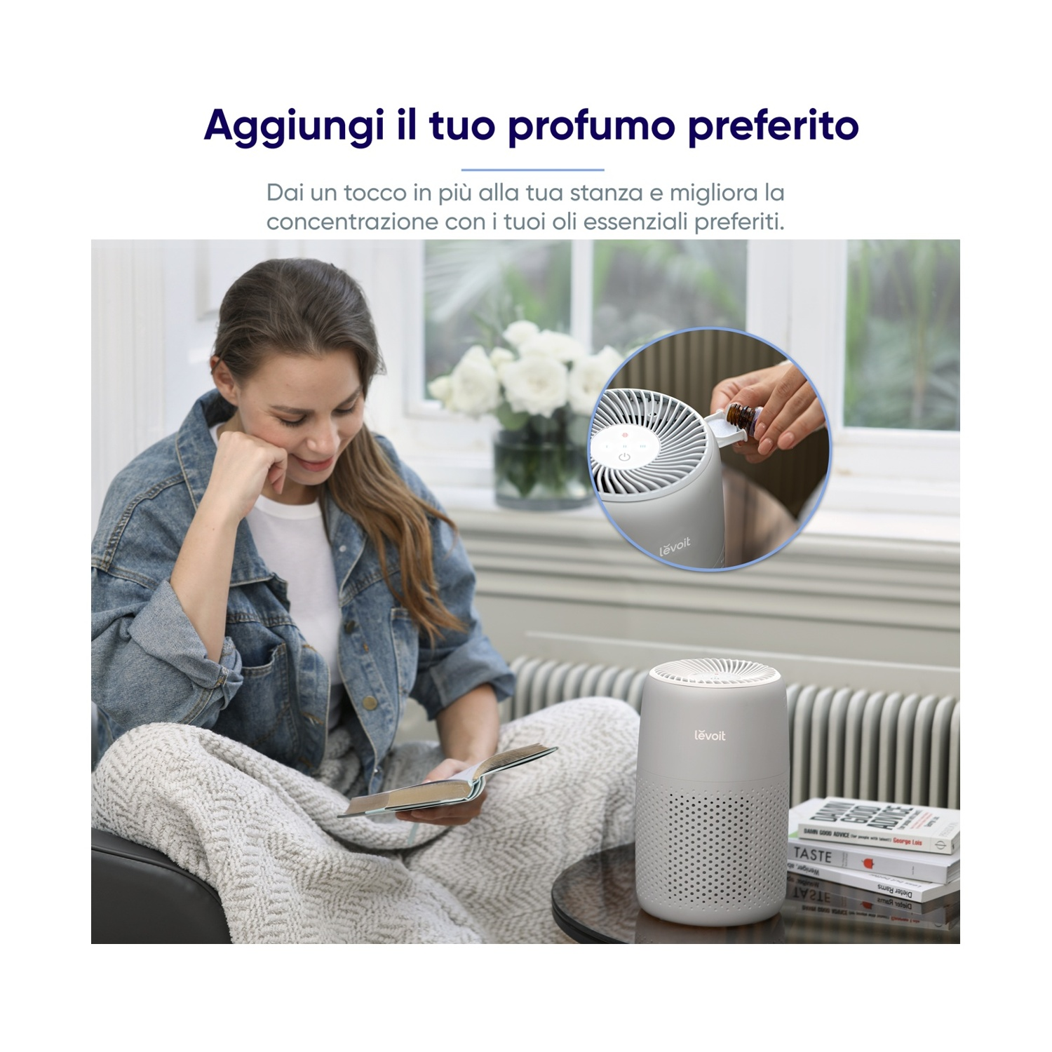 Donna in giacca di jeans seduta, che legge un libro, vicino a un purificatore d'aria grigio. Fiori bianchi su un tavolo e una persona che aggiunge olio.