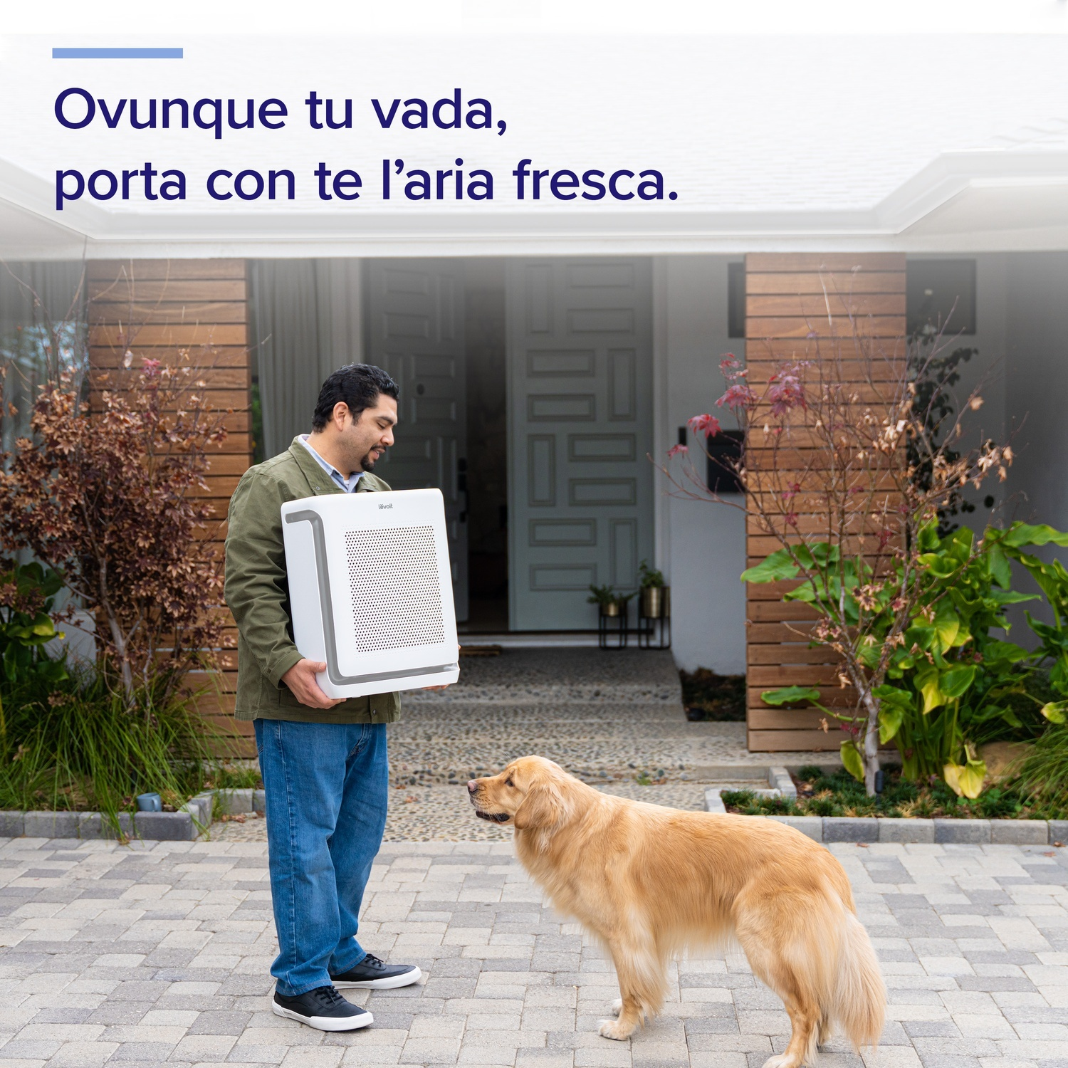 Un uomo tiene un purificatore d'aria, un golden retriever lo guarda. Sono su un sentiero di mattoni davanti a una casa.
