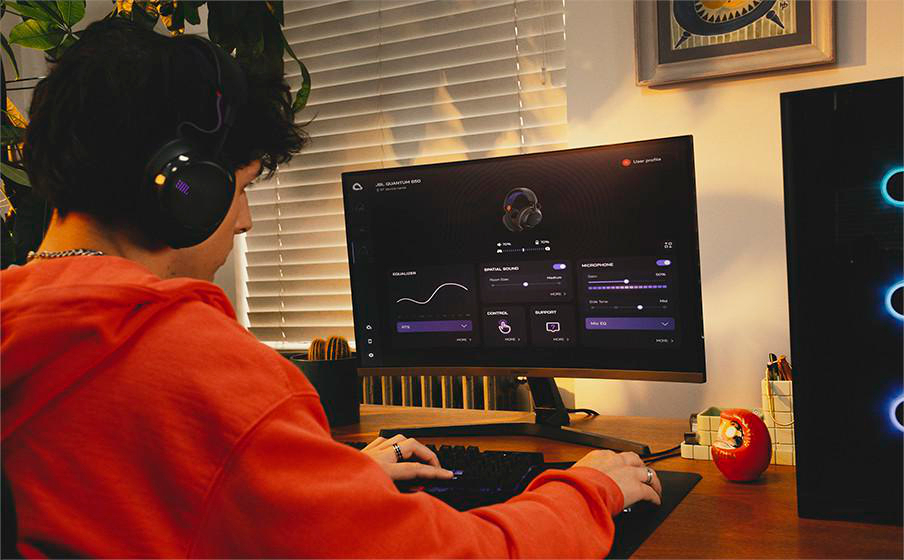 Eine Person mit Kopfhörern sitzt an einem Schreibtisch und blickt auf einen Monitor. Sie benutzt eine Tastatur. Rechts ist ein Computerturm.
