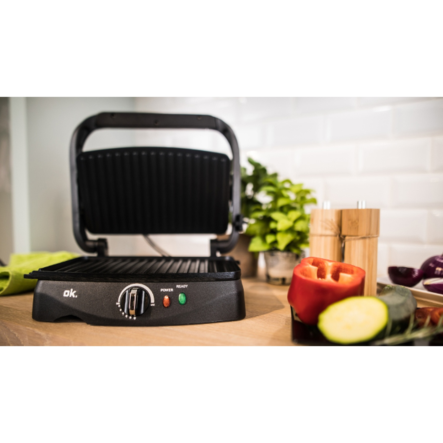 Una griglia nera aperta con verdure come peperoni rossi e zucchine sulla superficie di legno. Pianta verde sullo sfondo.