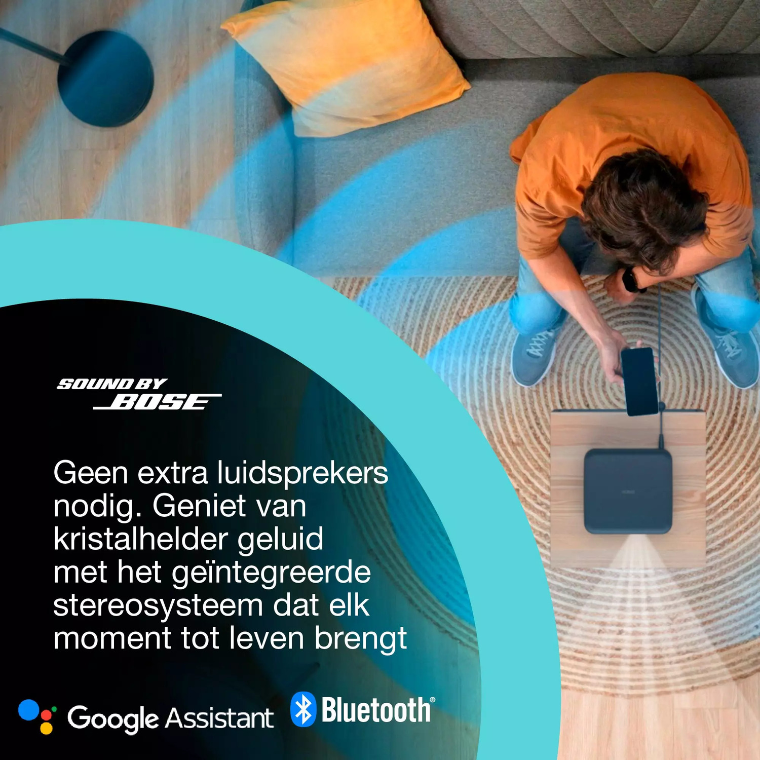 Een persoon zit met een smartphone die is aangesloten op een zwarte luidspreker. Een bank met een kussen staat op de achtergrond.