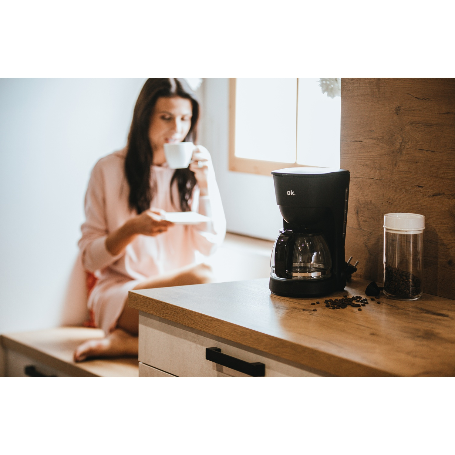 Una donna beve da una tazza, accanto a una macchina da caffè e un barattolo di fagioli su un bancone di legno. Chicchi di caffè sparsi.