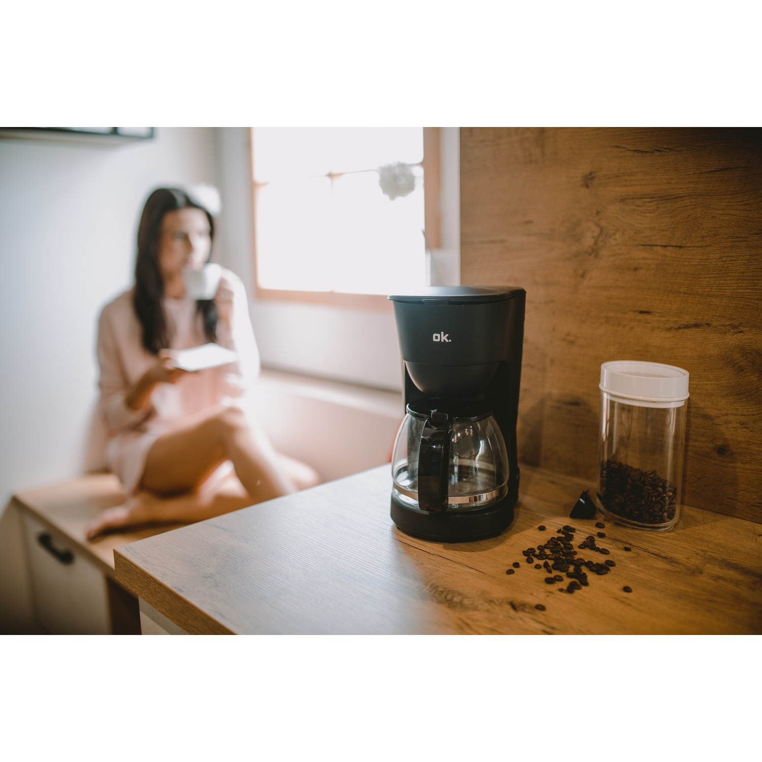 Una caffettiera nera, un barattolo con chicchi di caffè e chicchi sparsi su un bancone di legno. Una donna è sullo sfondo.