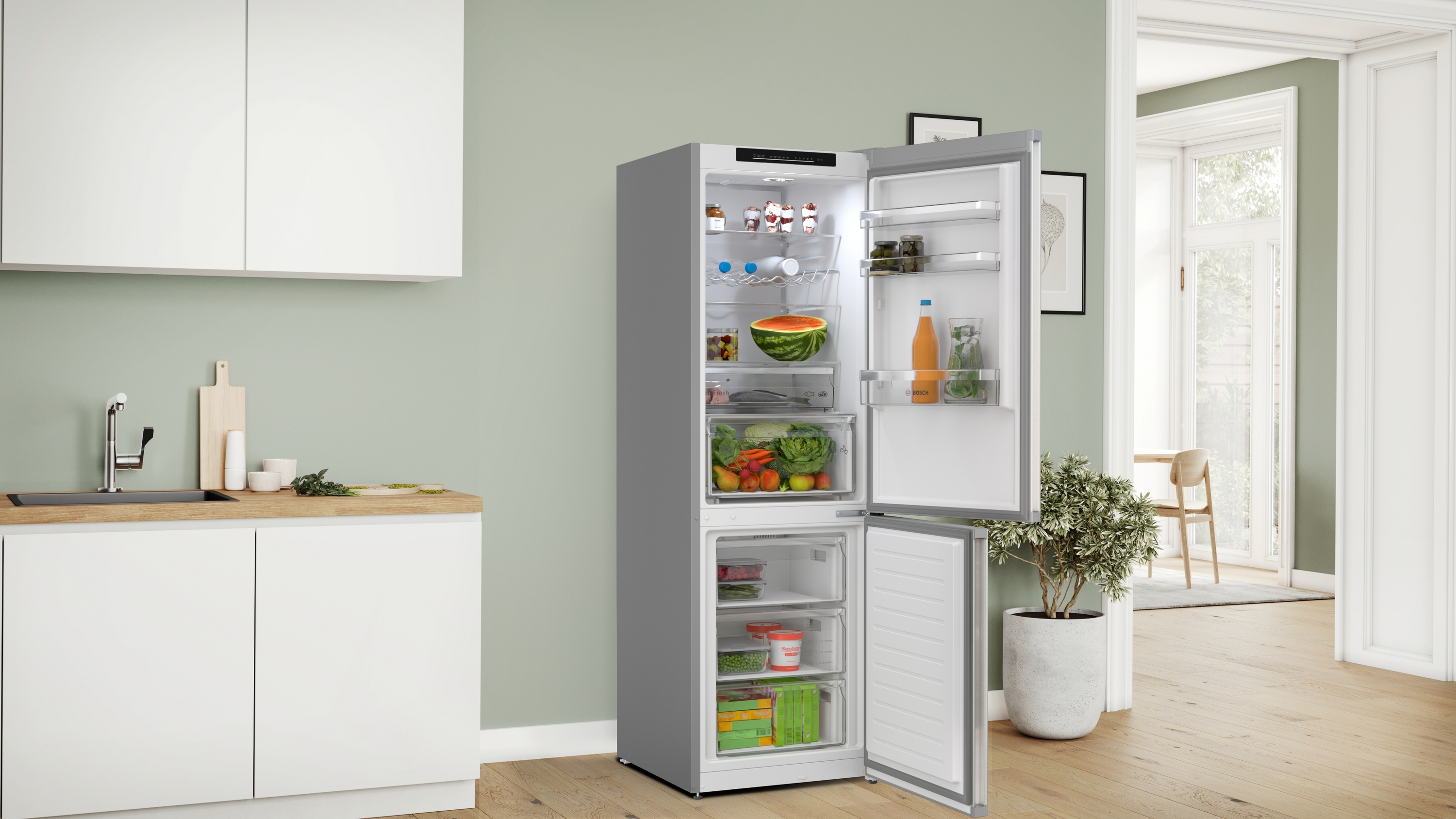 Offener Kühlschrank gefüllt mit Lebensmitteln. Küchenumgebung mit Schränken, Spüle und Tür. Eine Pflanze in einem Topf befindet sich auf der rechten Seite.