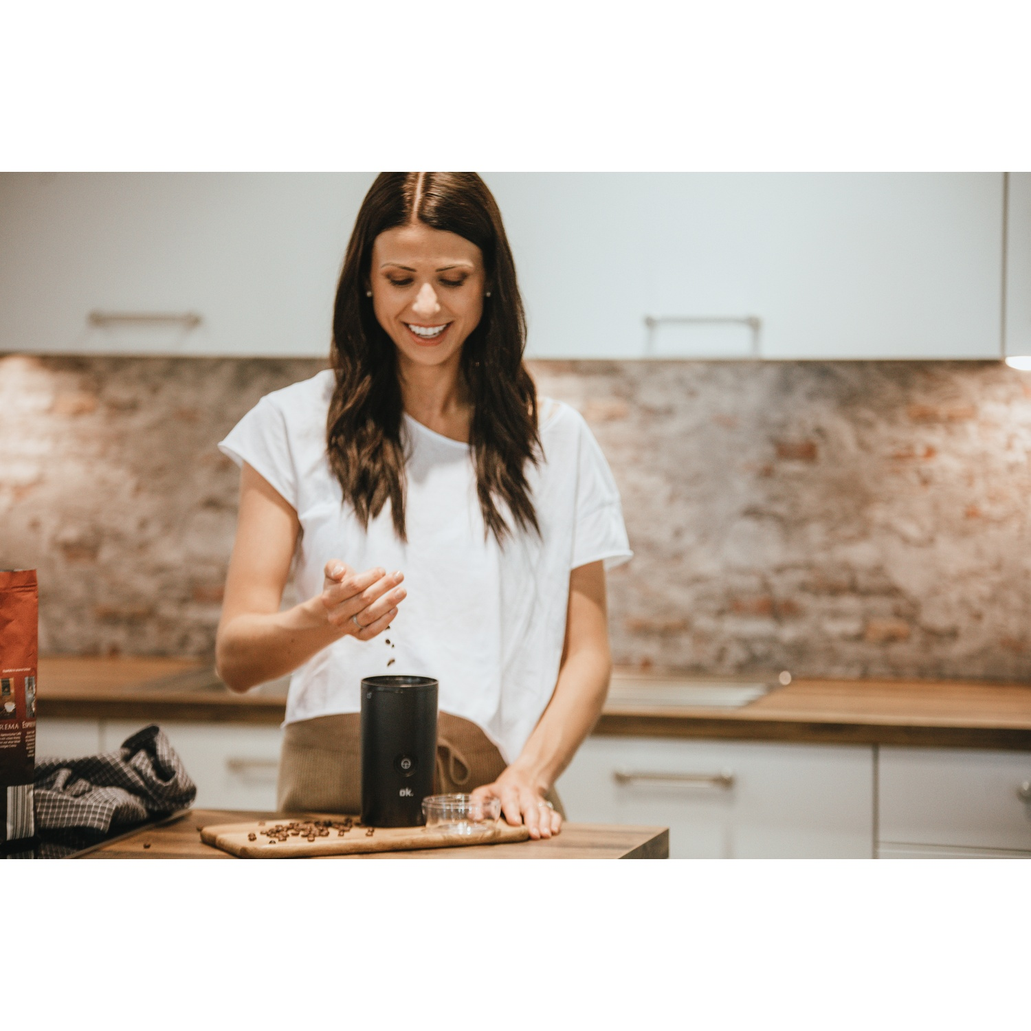 Una donna con una camicia bianca sorride, versando chicchi di caffè in un macinacaffè nero su un tagliere di legno in cucina.