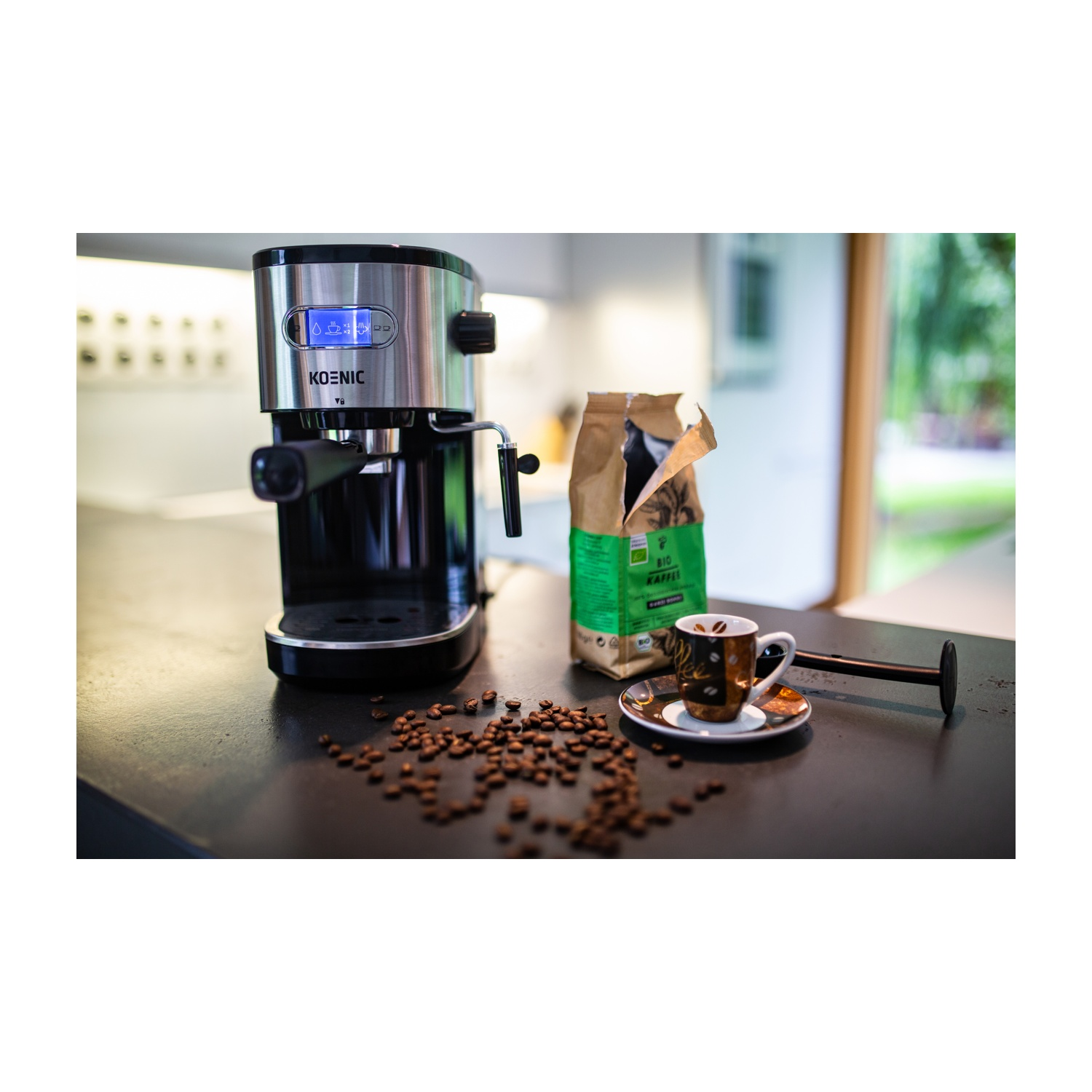 Macchina per caffè espresso, chicchi di caffè, sacchetto di caffè e tazza su un piano di lavoro nero. Lo sfondo è una cucina sfocata.