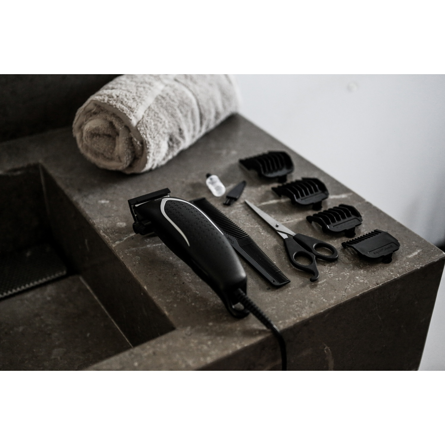 Un tagliacapelli nero, un pettine, delle forbici e diversi accessori sono su una superficie di pietra vicino a un asciugamano arrotolato.