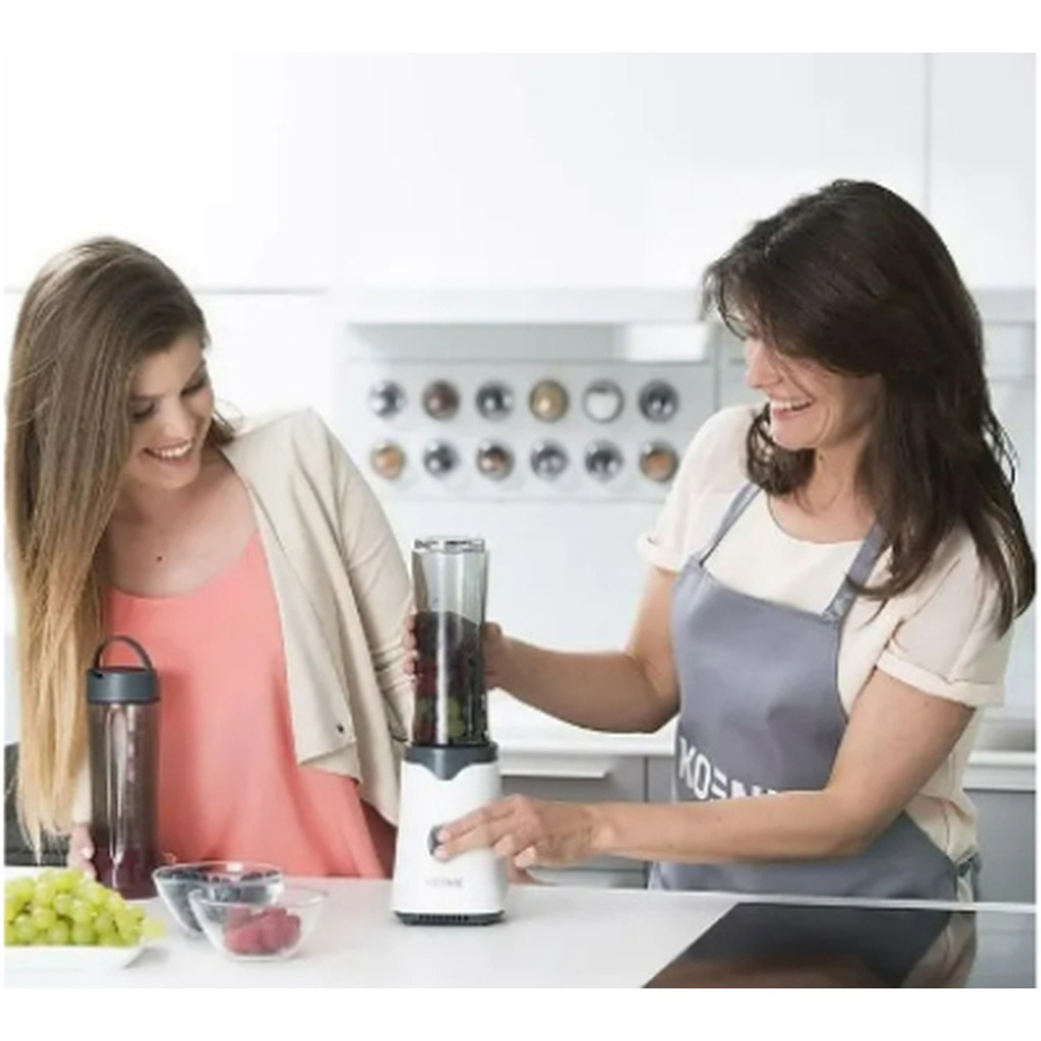 Due donne in cucina. Una frulla frutta in un frullatore, l'altra sorride. Uva, bacche e una bottiglia di frullato sono sul bancone.