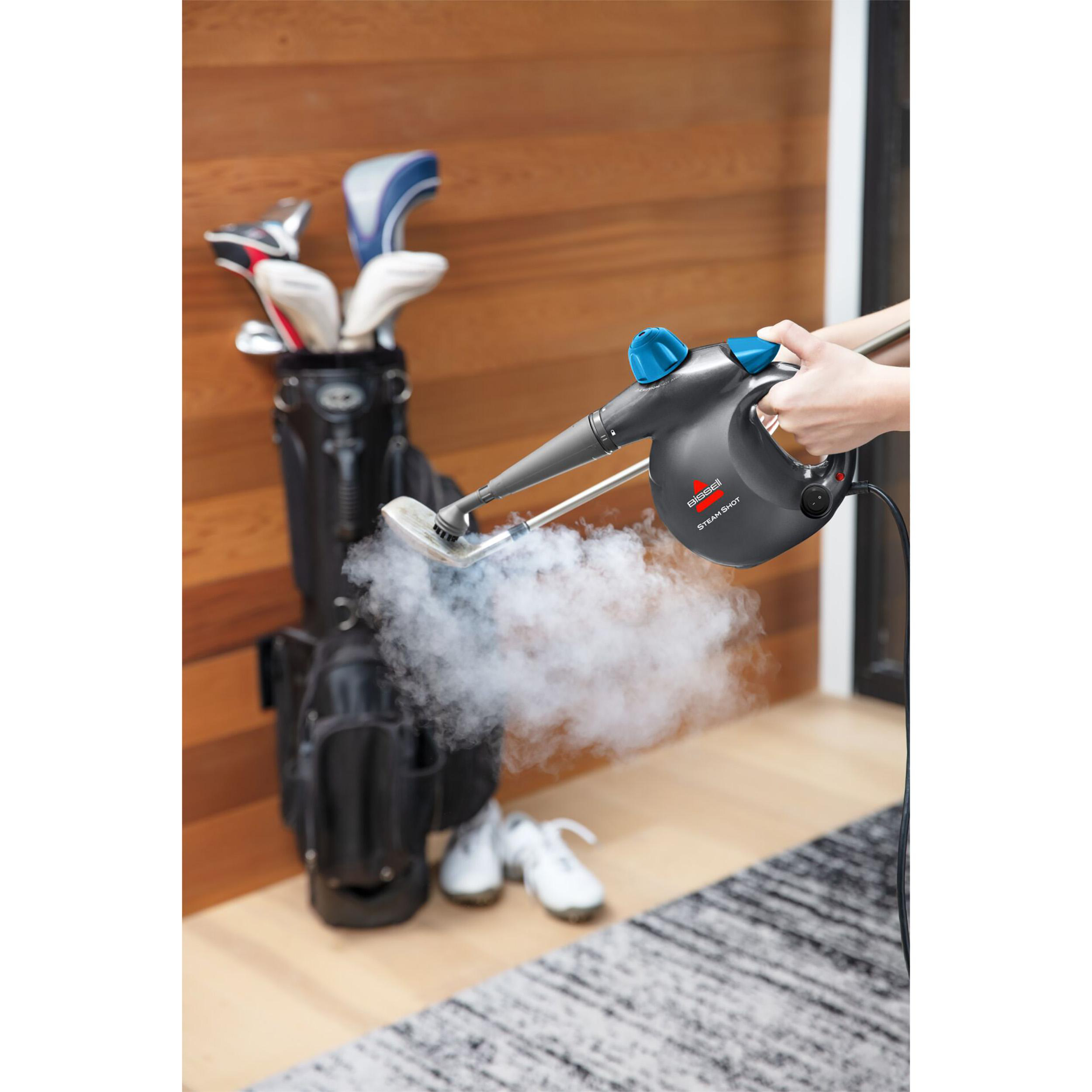 Eine Person reinigt einen Golfschläger mit einem Dampfreiniger. Eine Golftasche und Schuhe sind im Hintergrund sichtbar.