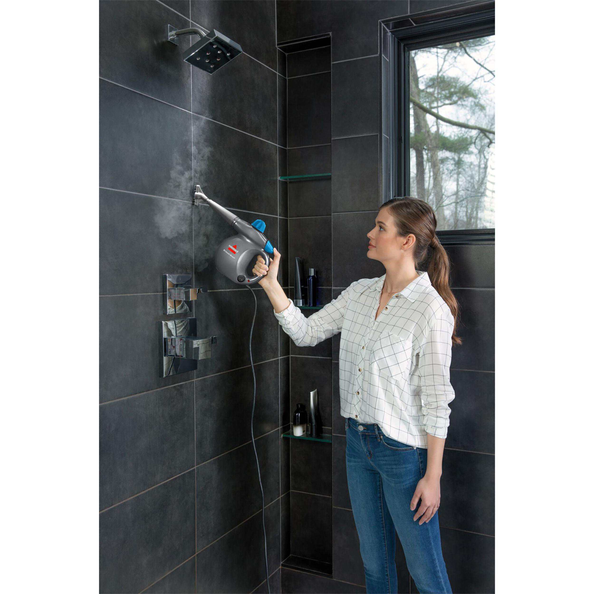 Eine Frau reinigt mit einem Dampfreiniger eine geflieste Dusche. Ein Fenster zeigt Bäume. Dampf ist sichtbar.