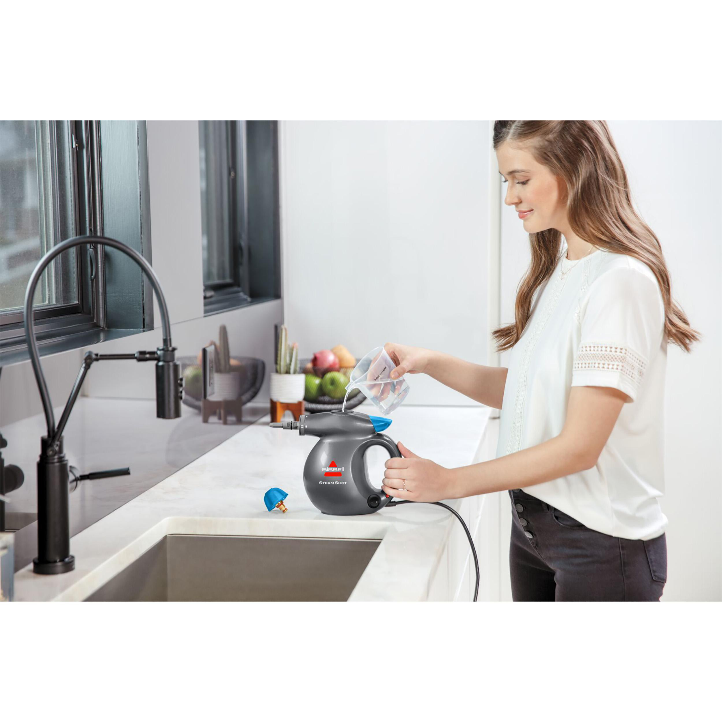 Eine Frau gießt Wasser in einen grauen Bissell Steam Shot. Die Szene ist eine Küche mit Spüle und Fenster.