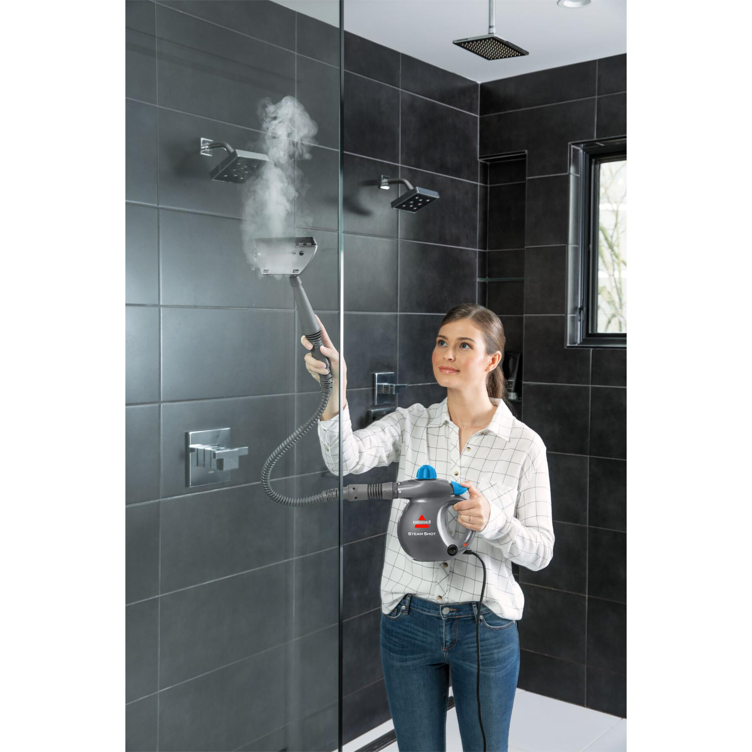 Eine Frau reinigt eine Dusche mit einem Dampfreiniger. Sie trägt Jeans und ein kariertes Hemd. Schwarze Fliesen sind im Hintergrund.