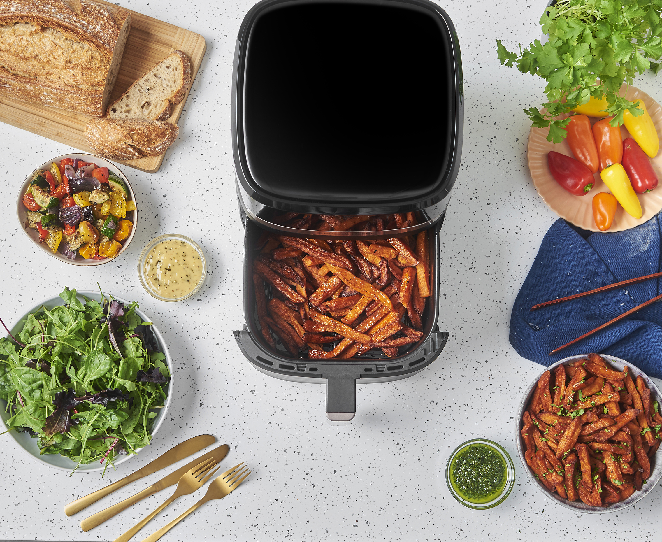 Felülnézet egy asztalról, amelyen egy airfryer, ételek, például sült krumpli, kenyér, paprika, saláta és szószok találhatók.