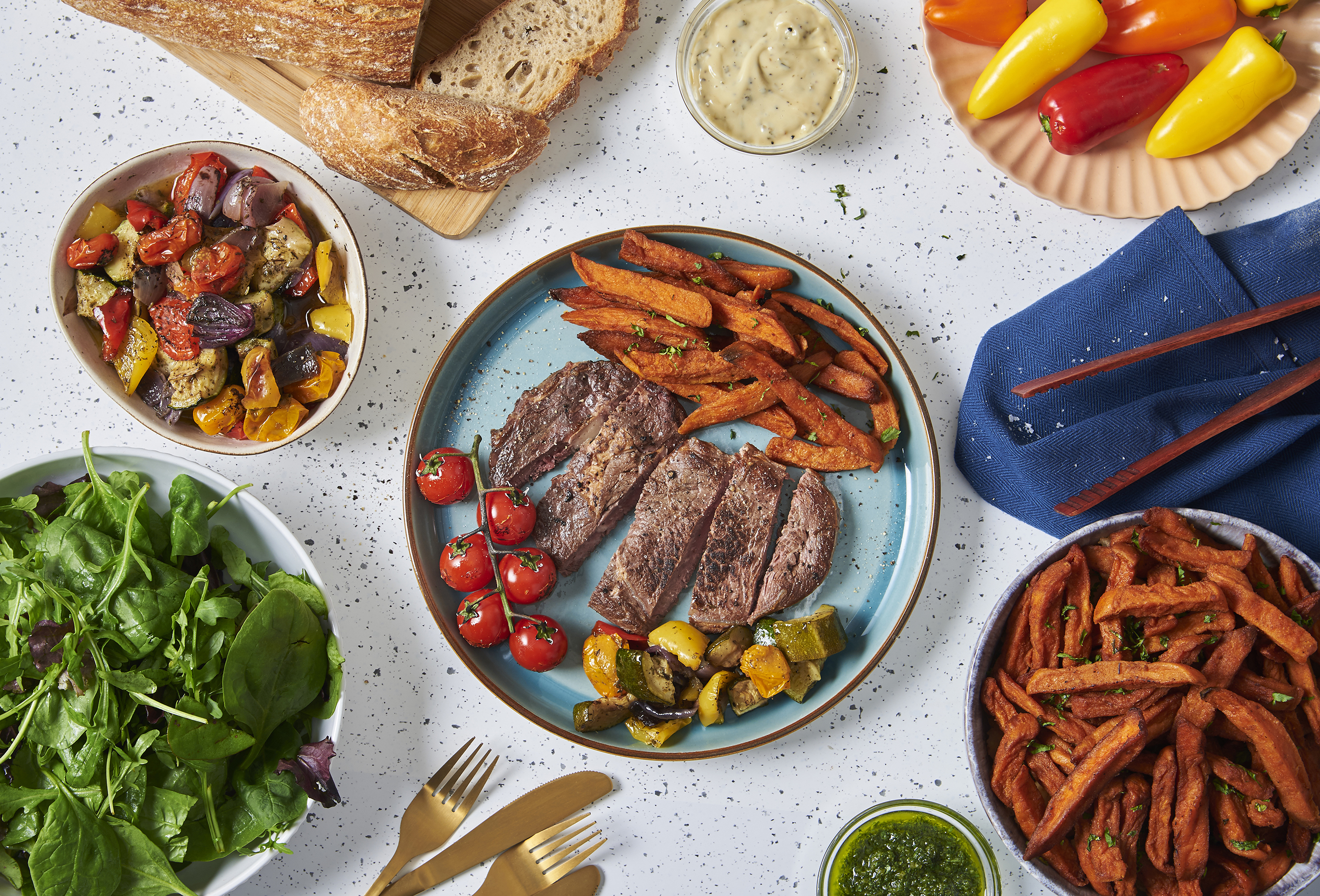 Felülnézet egy étkezésről. Steak, édesburgonya sült krumpli, zöldségek, kenyér és paprika fehér felületen elrendezve.