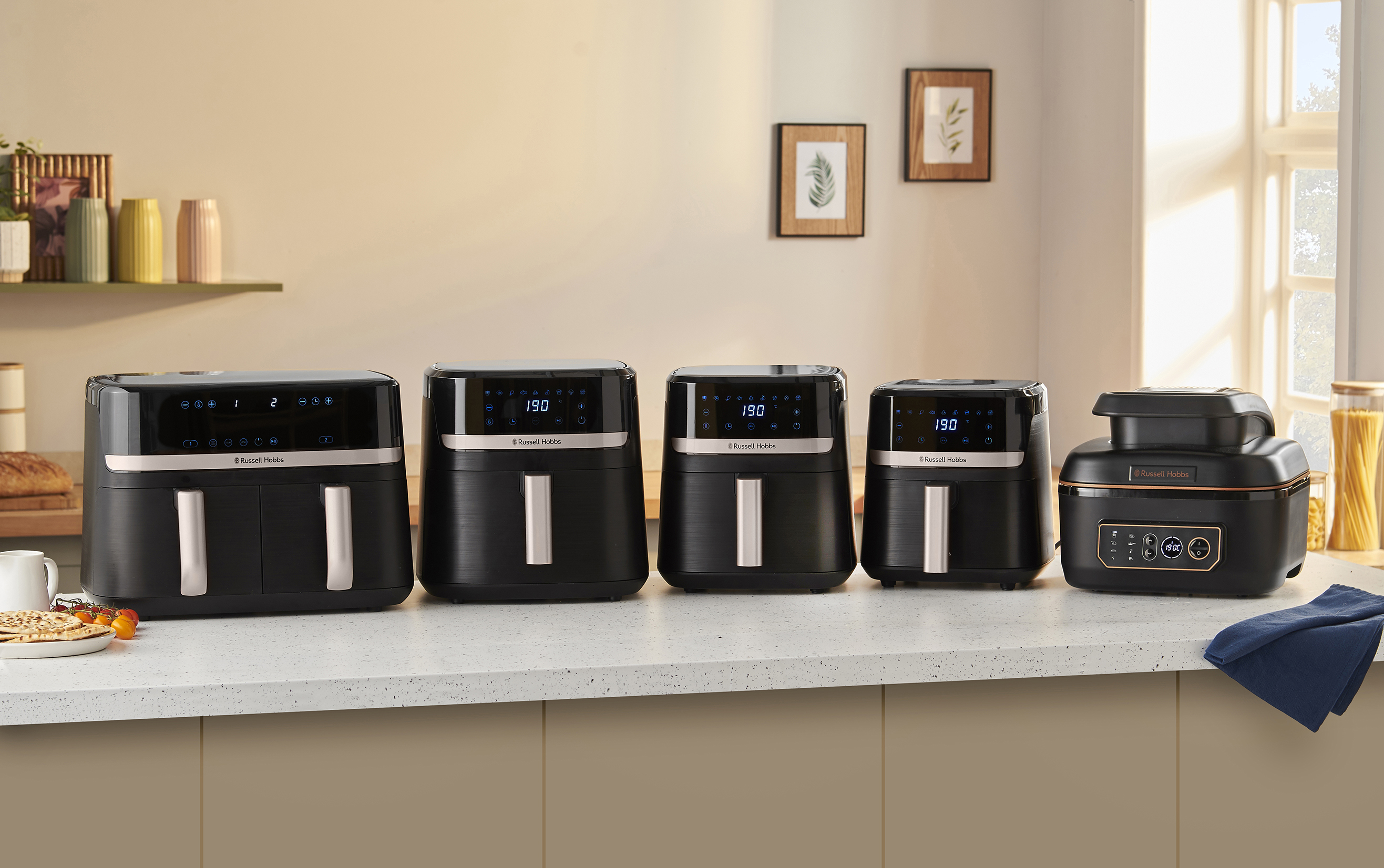 Több fekete Russell Hobbs légkeveréses fritőz különböző méretben egy pulton. Egy ablak és egy polc dekorációval a háttérben.