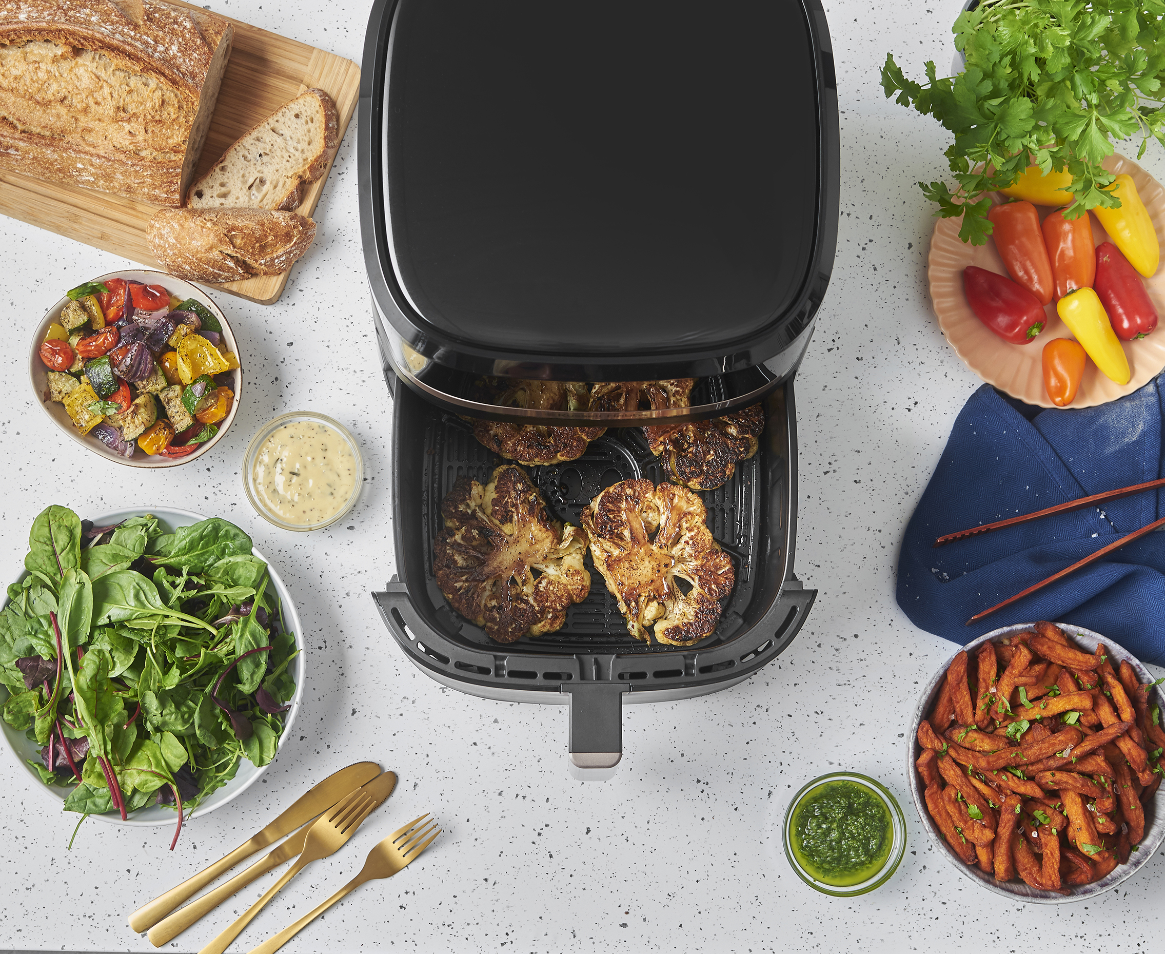 Felülnézet egy air fryer-ről, sült karfiollal, salátákkal, kenyérrel, zöldségekkel és szószokkal körülvéve.