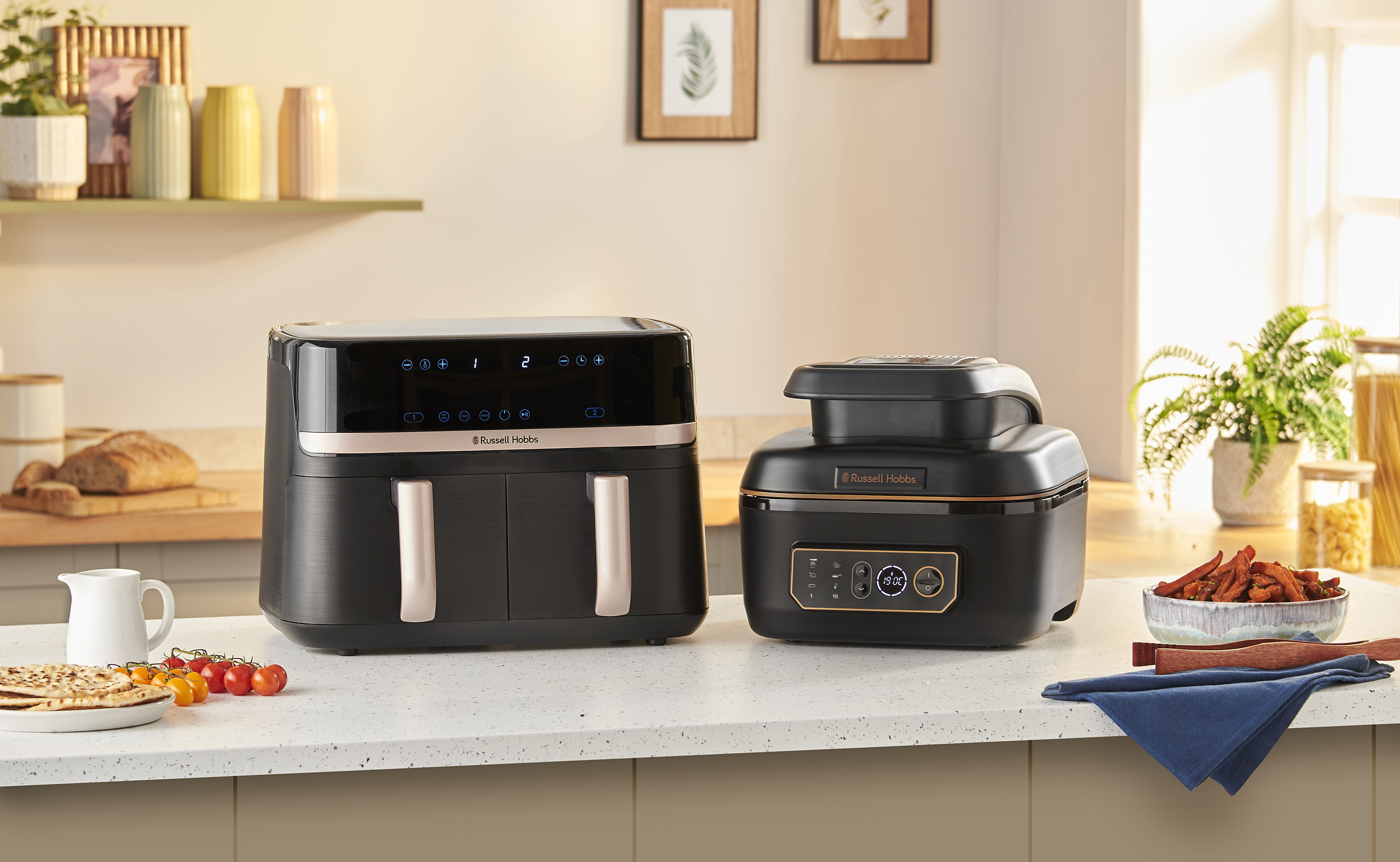 Két Russell Hobbs légkeveréses fritőz a pulton, élelmiszerekkel. A háttérben növények és polcok láthatók.