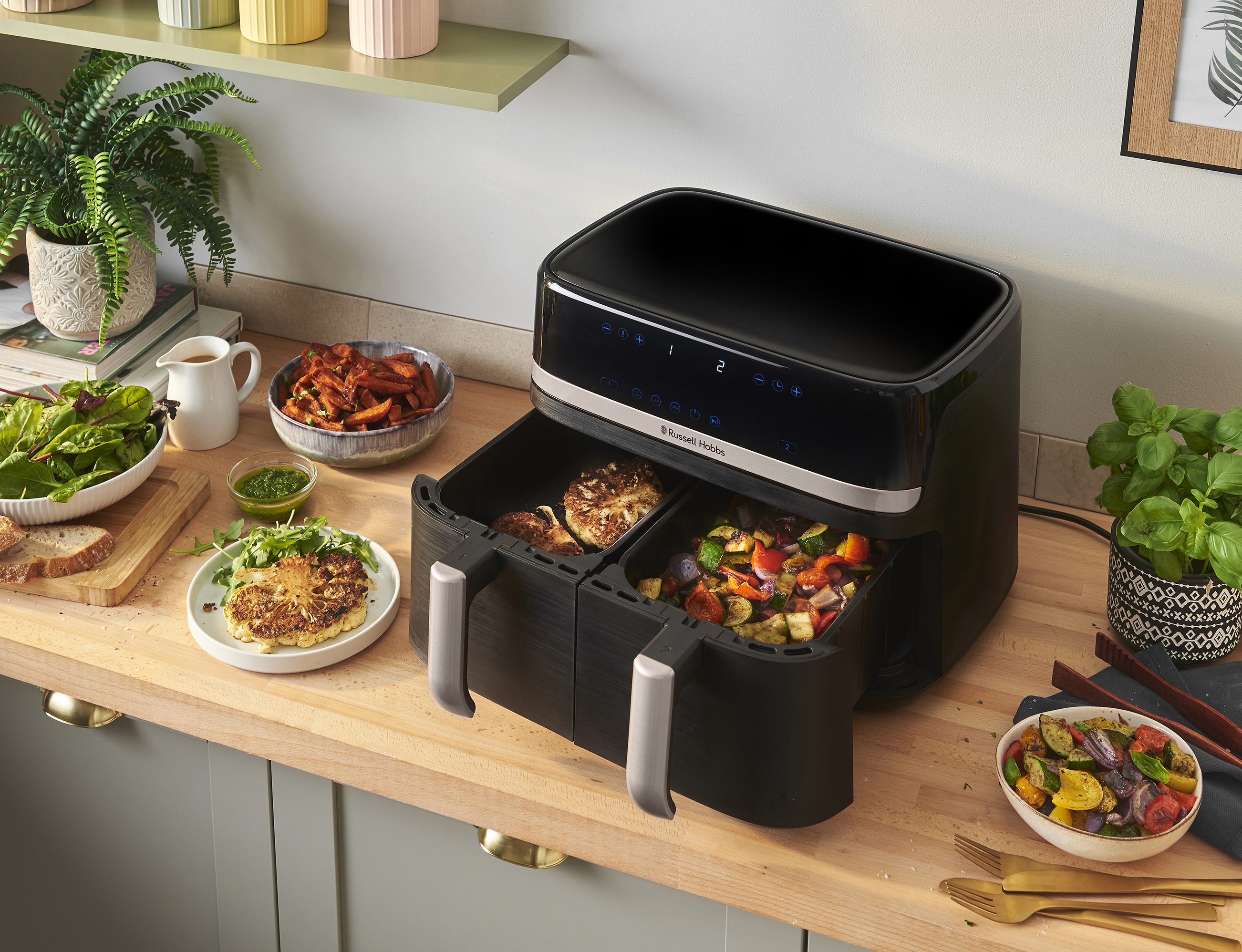 Egy airfryer főtt étellel, zöldségekkel és salátával a fa pulton. Egy növény és más ételek is láthatók.