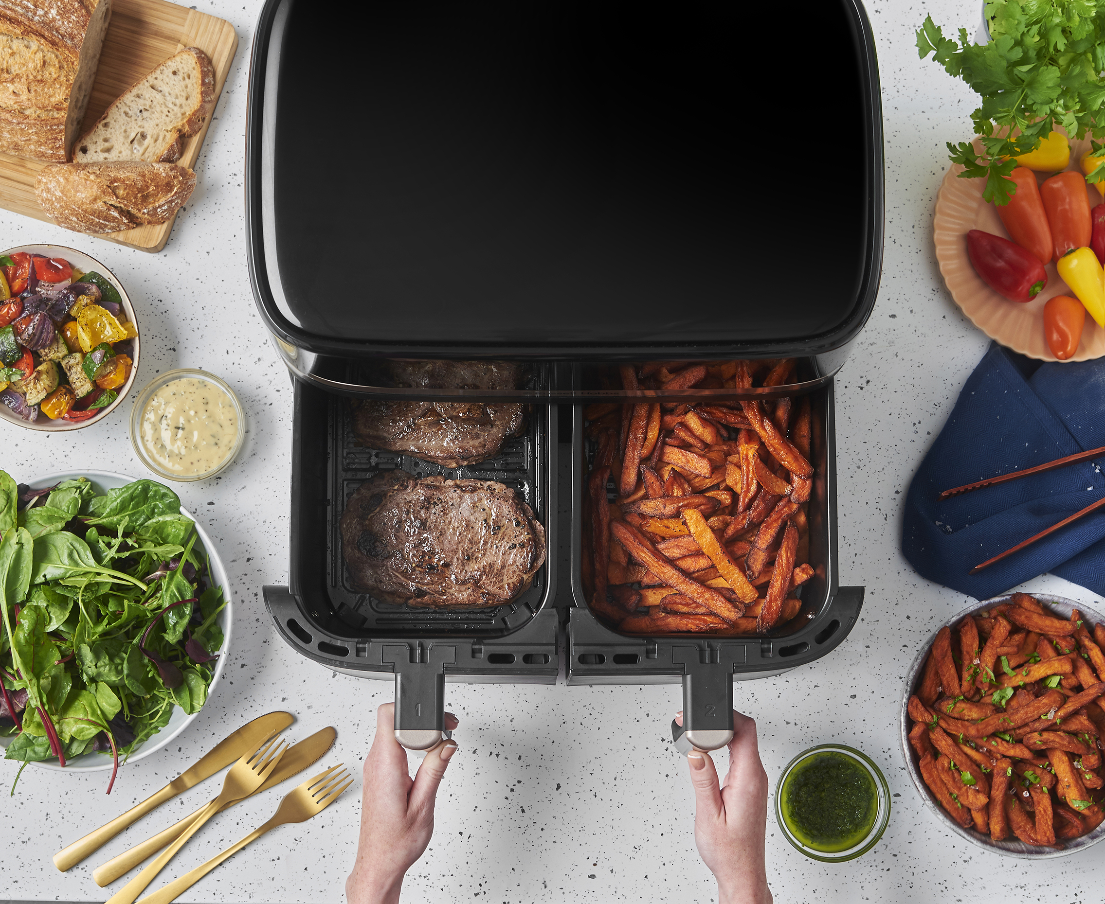 Felülnézet a légsütőről steakkel és sült krumplival, salátával, kenyérrel, zöldségekkel, szószokkal körülvéve, kezekkel, amik a sütőt tartják.
