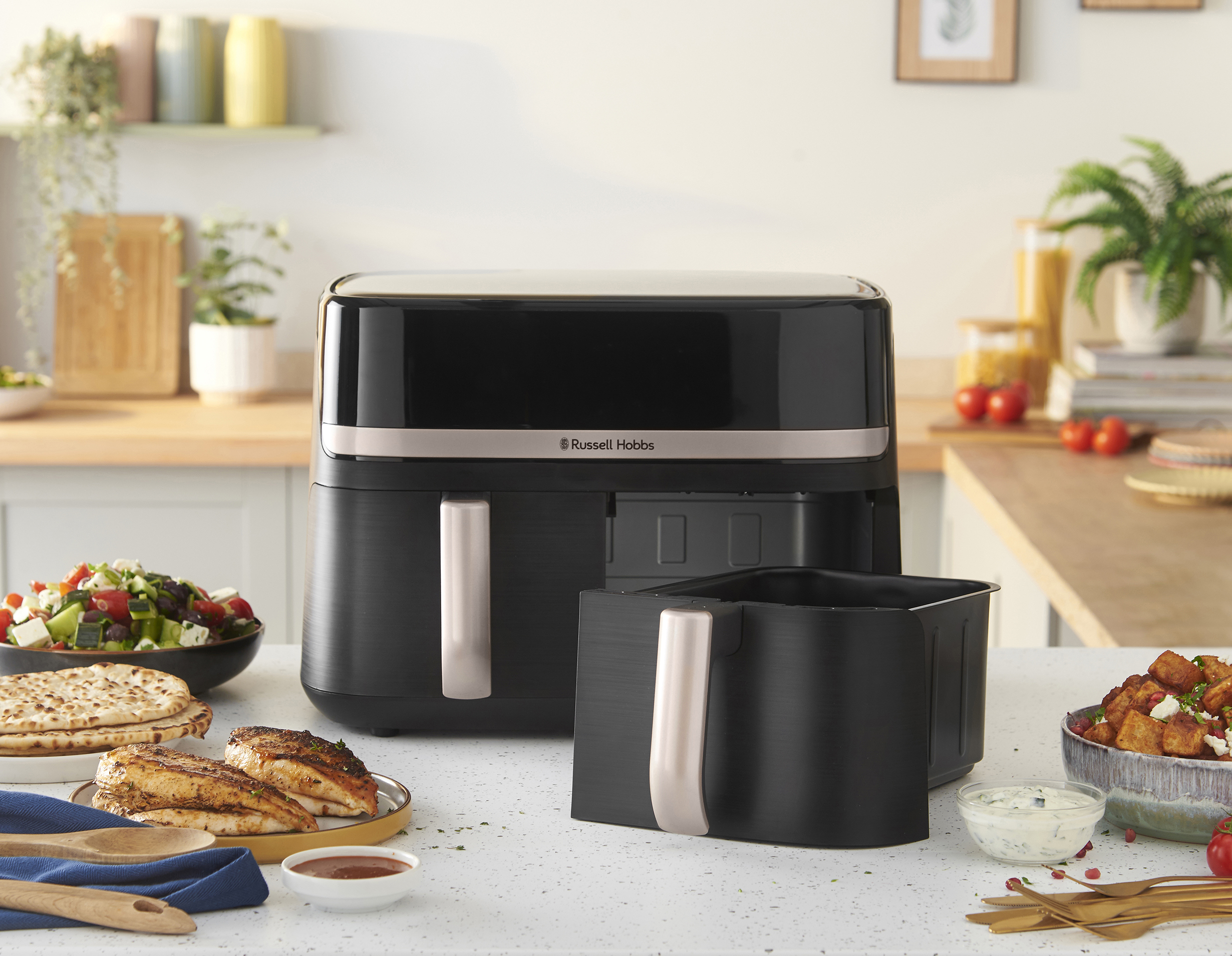 Fekete légsütő konyhapulton étellel. Saláta, csirke és burgonya látható. Russell Hobbs márka.