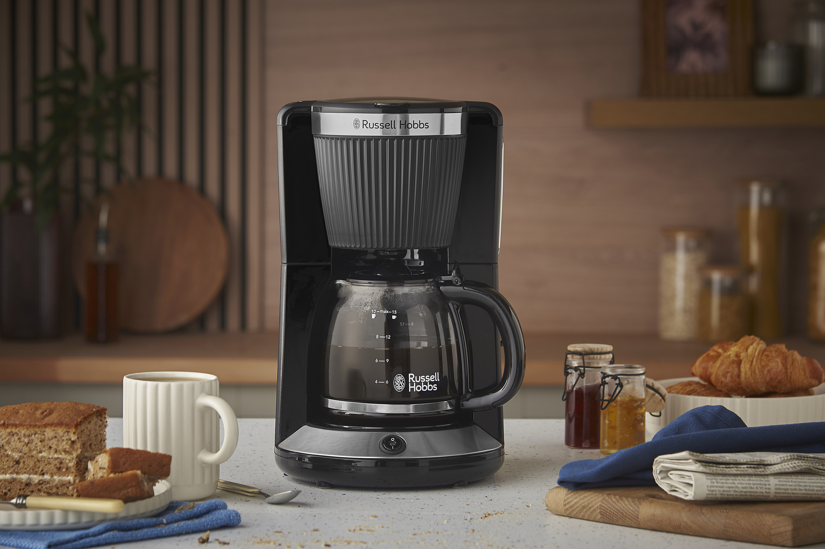 Egy fekete Russell Hobbs kávéfőző a pulton. Mellette sütemény, bögre és croissant látható.
