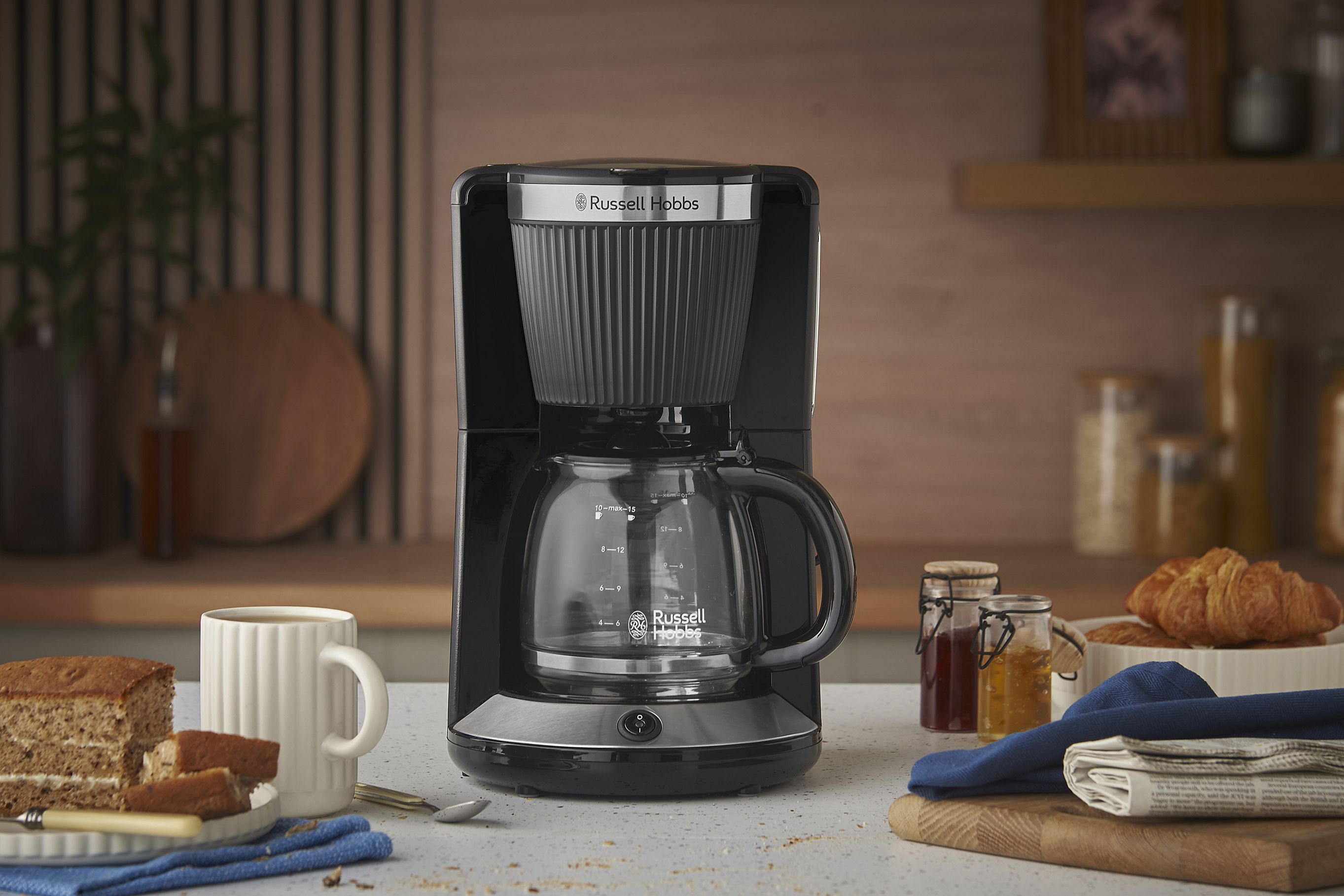 Fekete Russell Hobbs kávéfőző. Sütemény, bögre, croissant és lekváros üvegek a pulton.