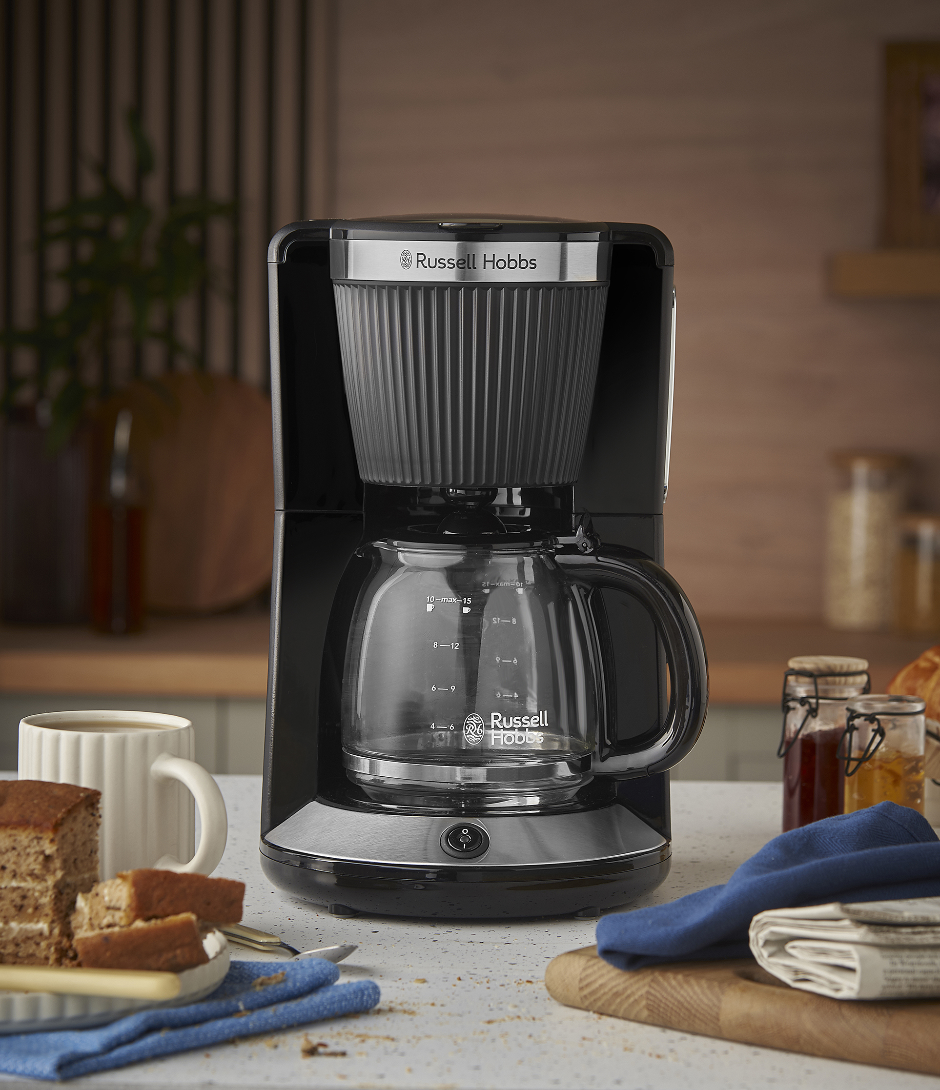 Egy fekete Russell Hobbs kávéfőző a pulton süteménnyel, kávéval és lekváros üvegekkel. Egy kék kendő és egy újság van a közelben.