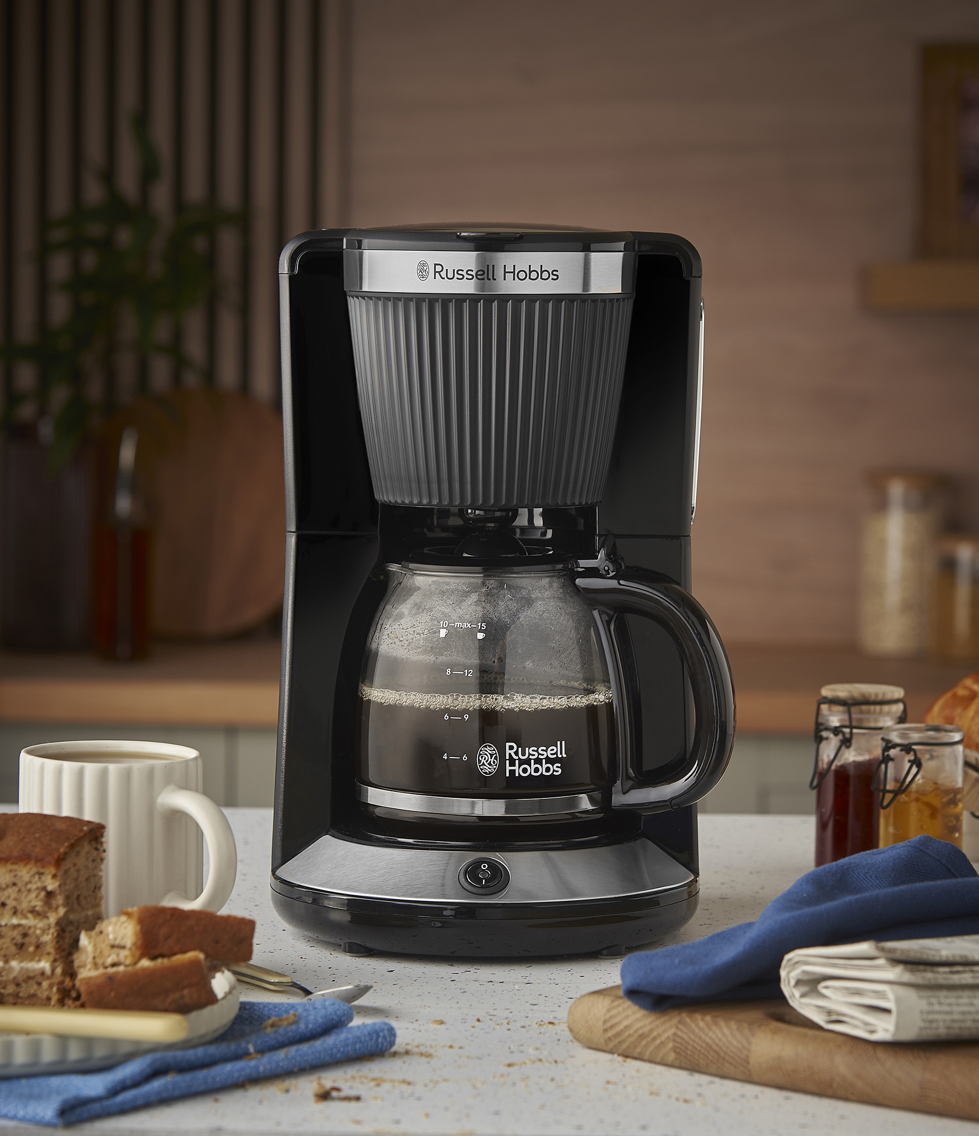 Egy fekete Russell Hobbs kávéfőző kávéval, süteménnyel és bögrével. A háttérben lekváros üvegek és egy újság.