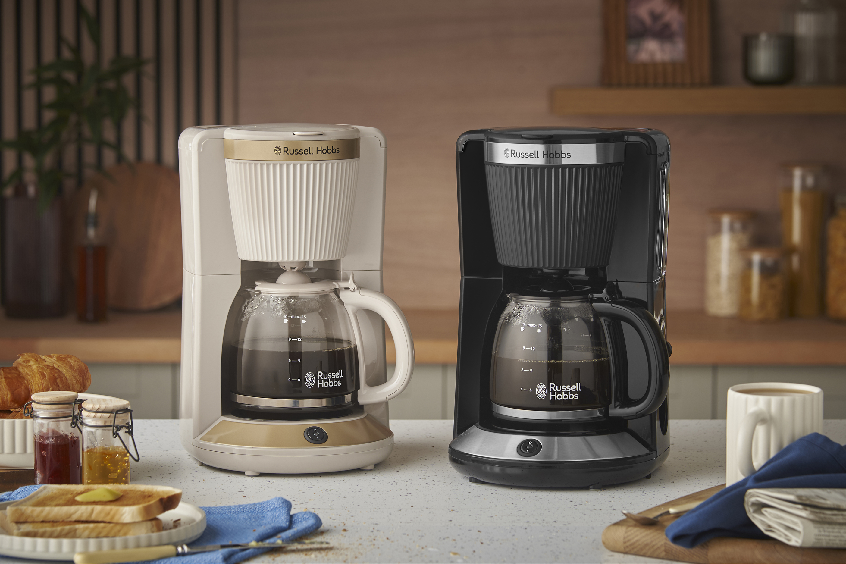 Két Russell Hobbs kávéfőző, egy krémszínű és egy fekete, pulton reggeli ételekkel.