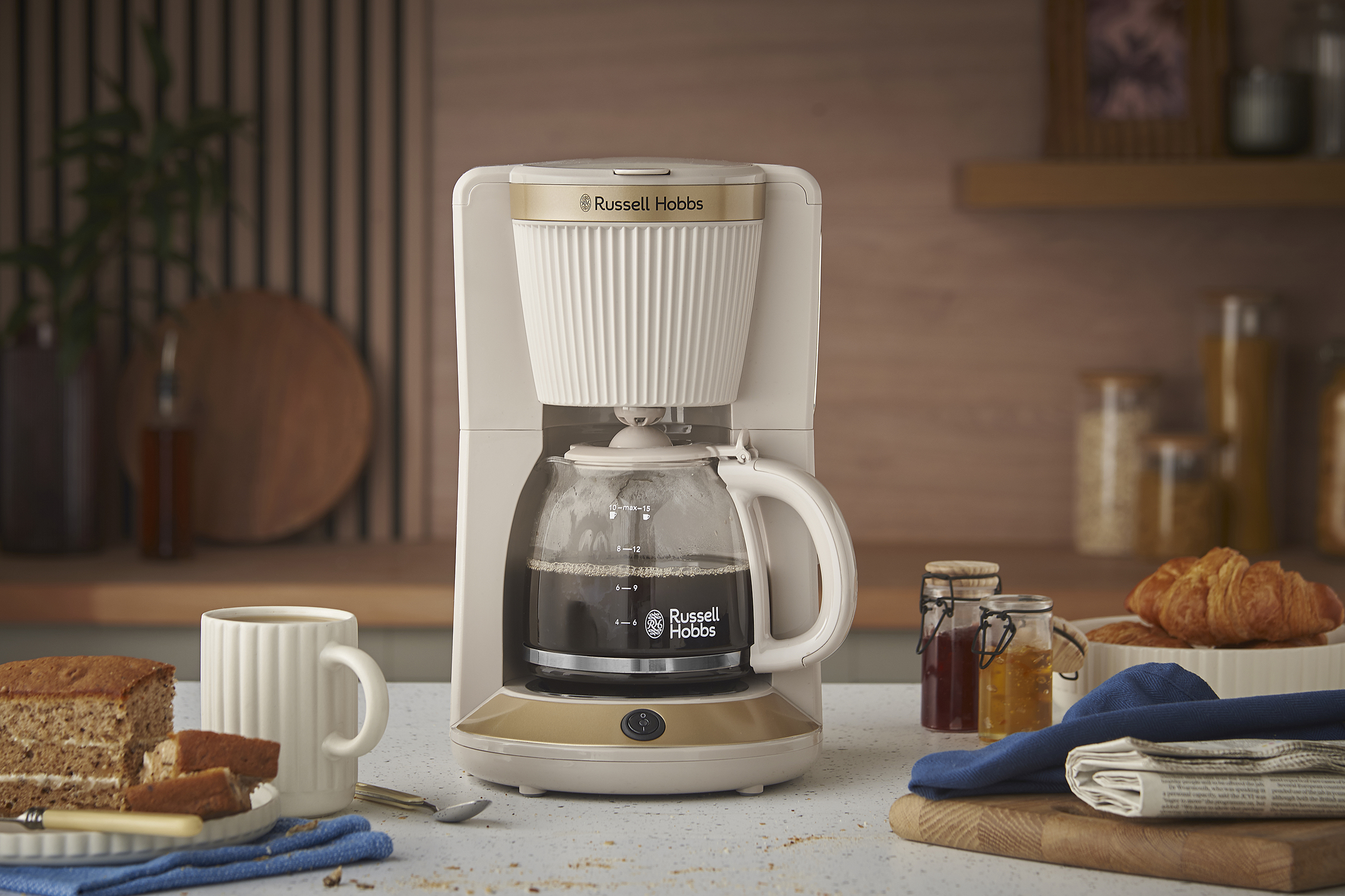Egy Russell Hobbs kávéfőző üvegkancsóval, süteménnyel, bögrével és croissant-okkal az asztalon.