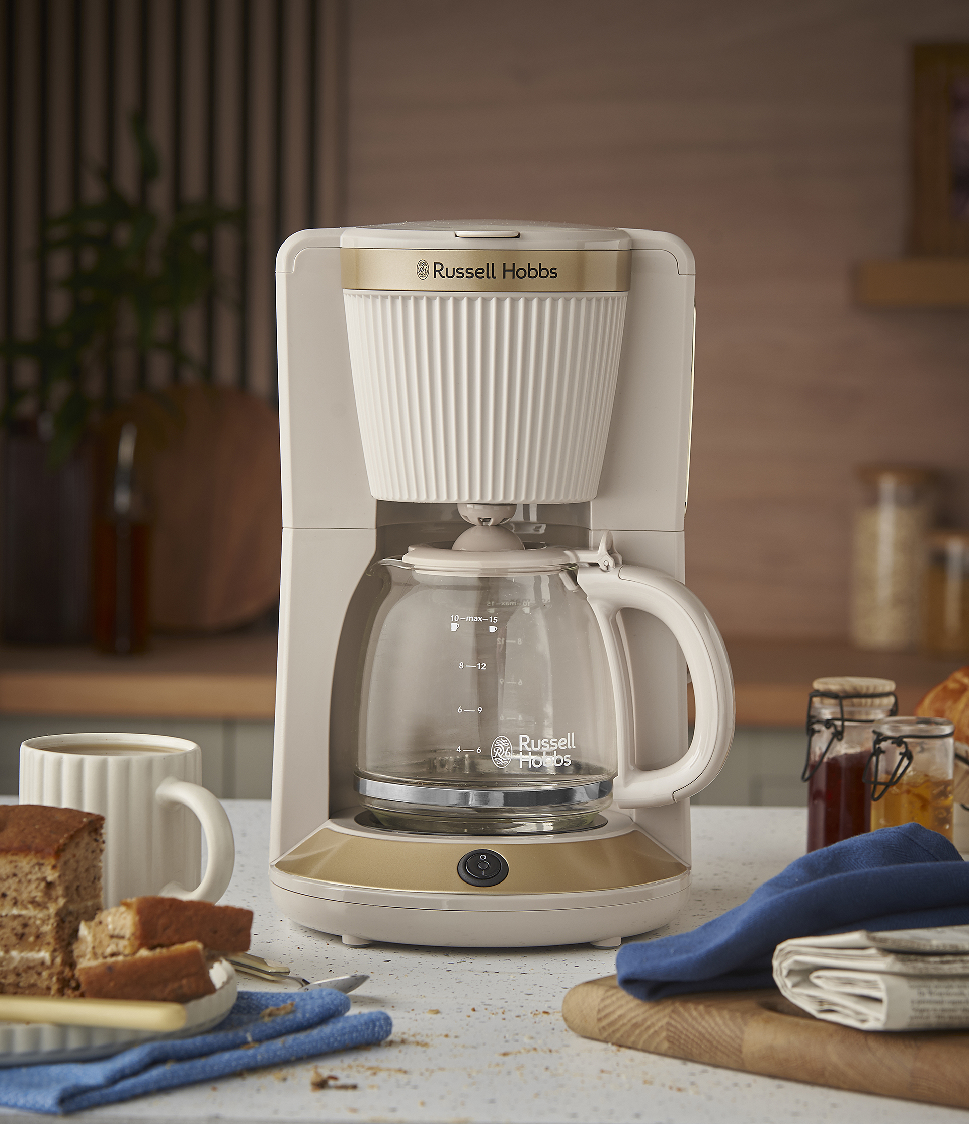 Egy Russell Hobbs kávéfőző a pulton, süteménnyel és bögrével. Üvegek, fadarab és újság a háttérben.