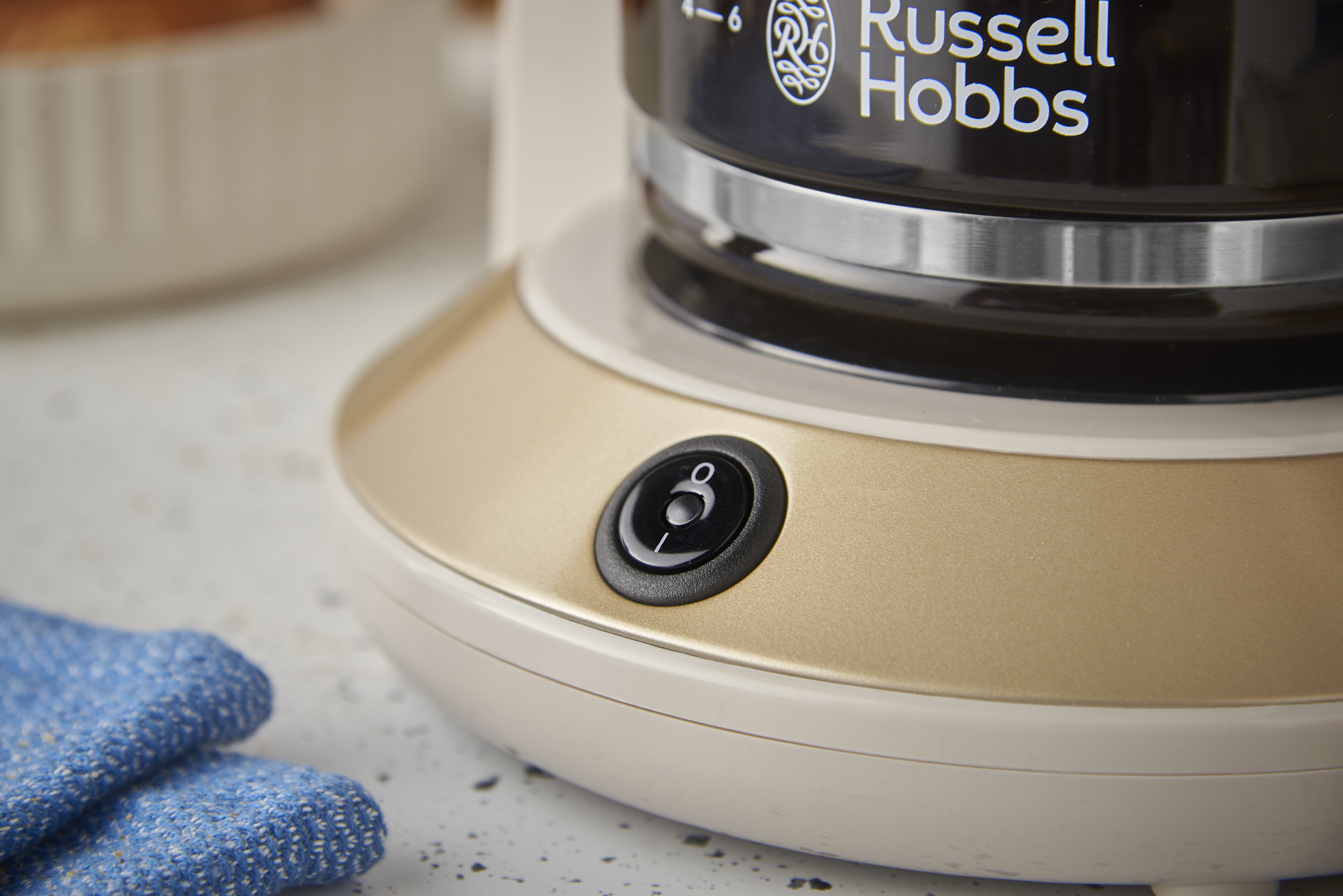 Egy Russell Hobbs kávéfőző bekapcsológombot és az üvegkancsó egy részét mutatja. Egy kék rongy látható.