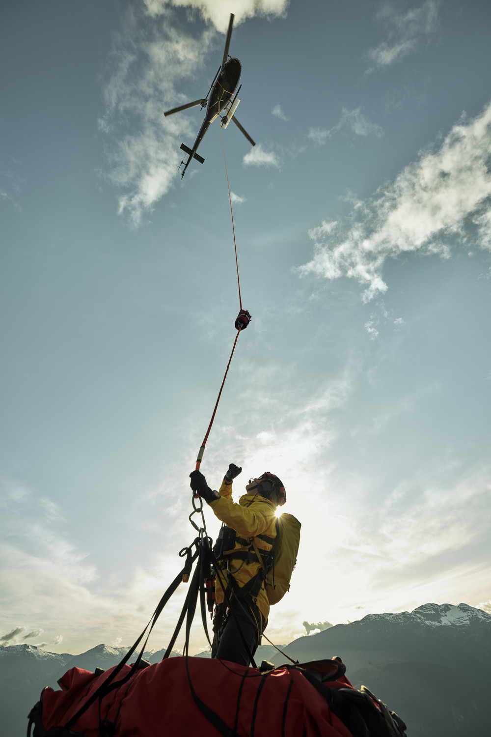 Un helicóptero rescata a una persona. La persona lleva equipo amarillo y está atada a una cuerda. Montañas y cielo en el fondo.