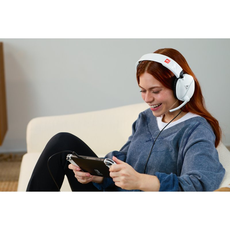 Una mujer pelirroja con auriculares sonríe mientras juega un juego portátil. Está sentada en un sofá blanco.