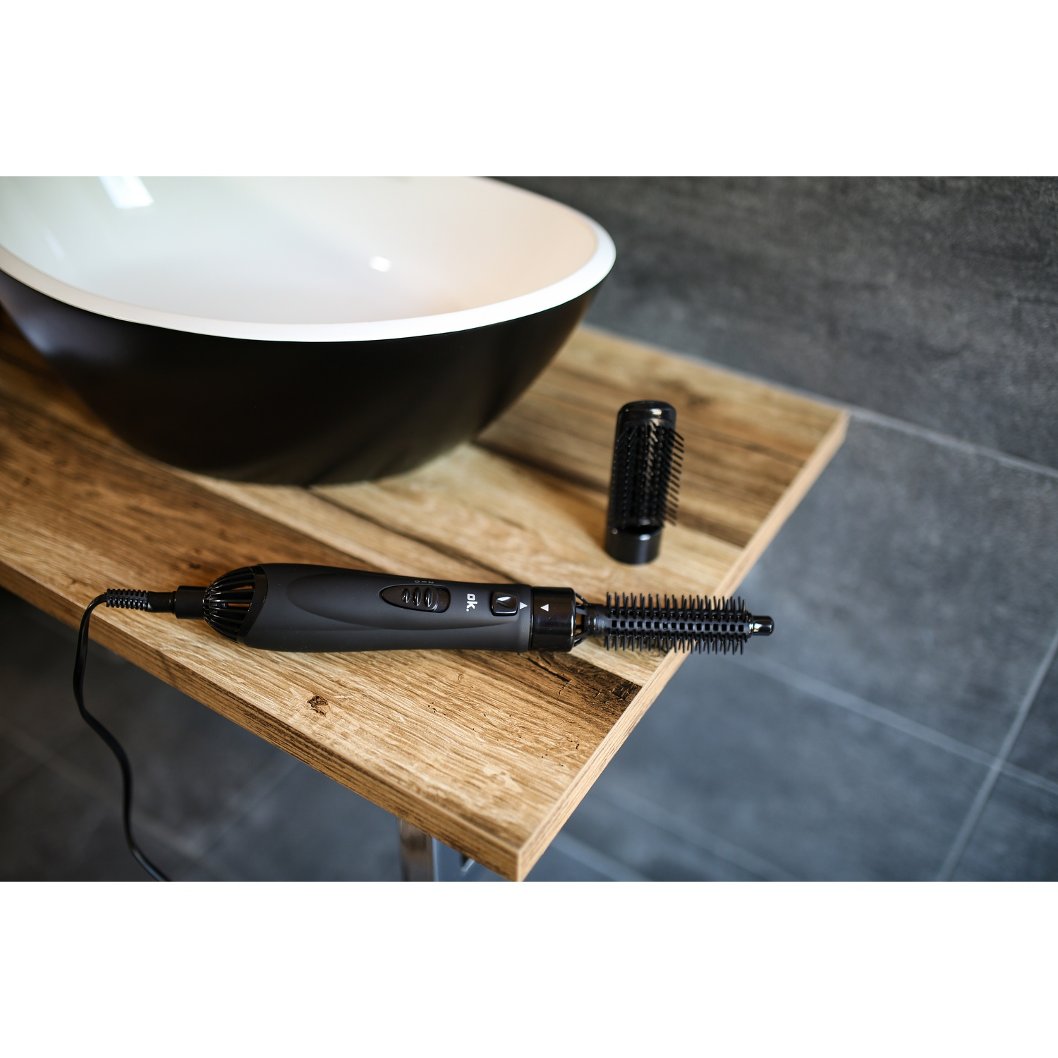 Un arricciacapelli nero con accessori poggia su una mensola di legno. Un lavabo bianco e nero sullo sfondo.