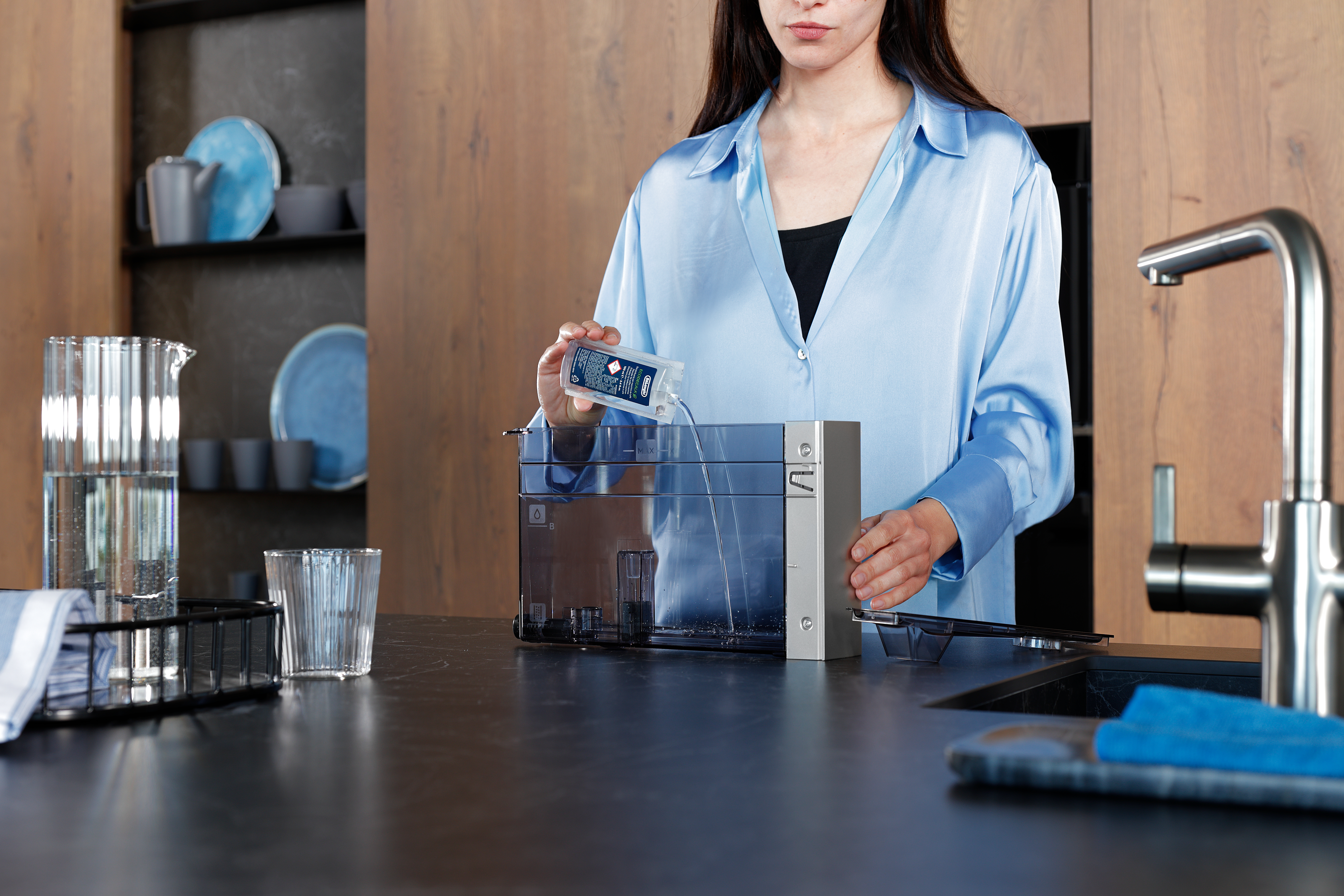 Eine Frau gießt Wasser aus einem Filter in eine Maschine. Eine Kanne, ein Glas und ein Wasserhahn sind ebenfalls in der Küche zu sehen.